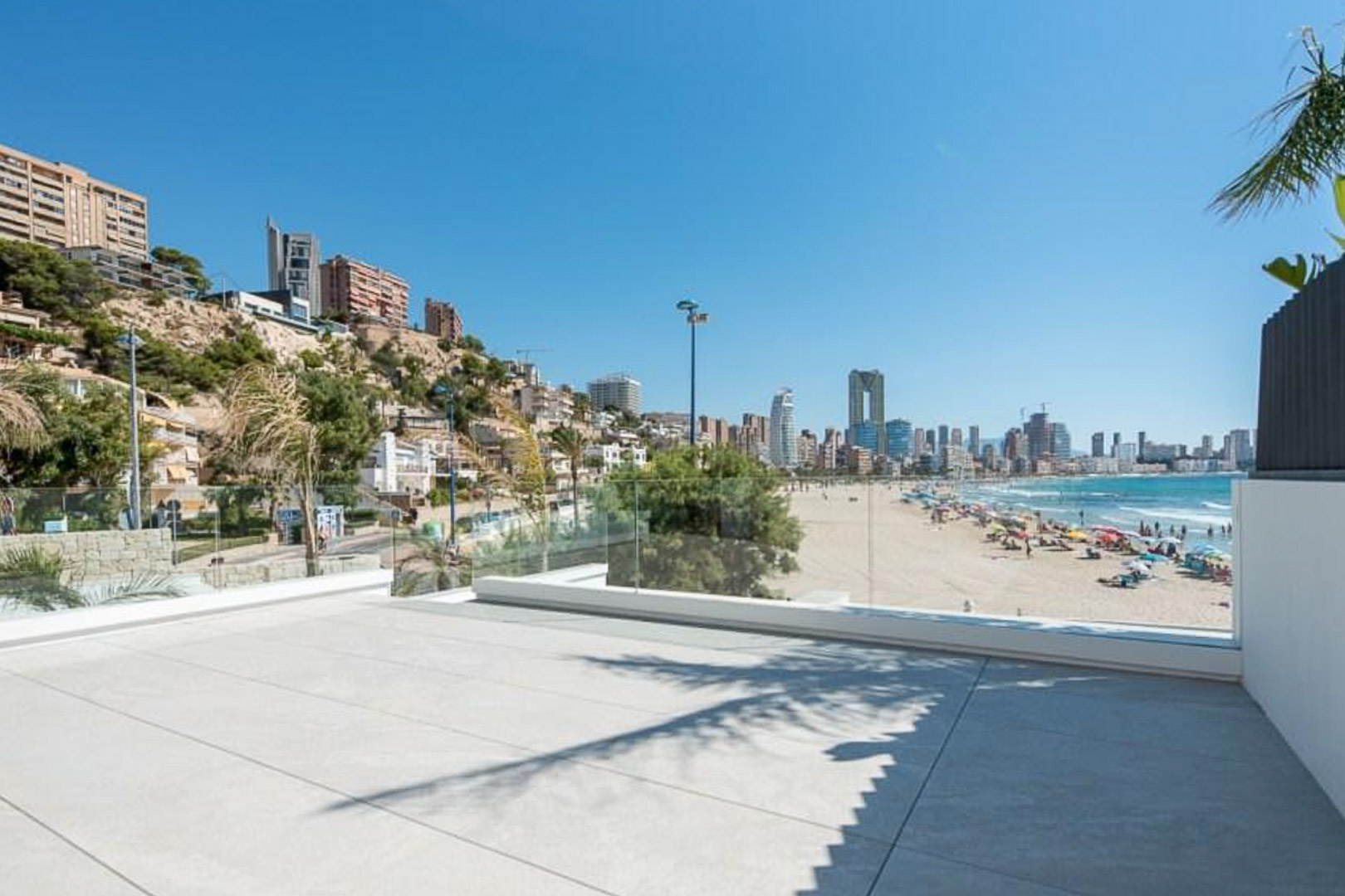 Buitenterras met grootformaat tegels en glazen balustrades, met panoramisch uitzicht over het zandstrand en de skyline van Benidorm.