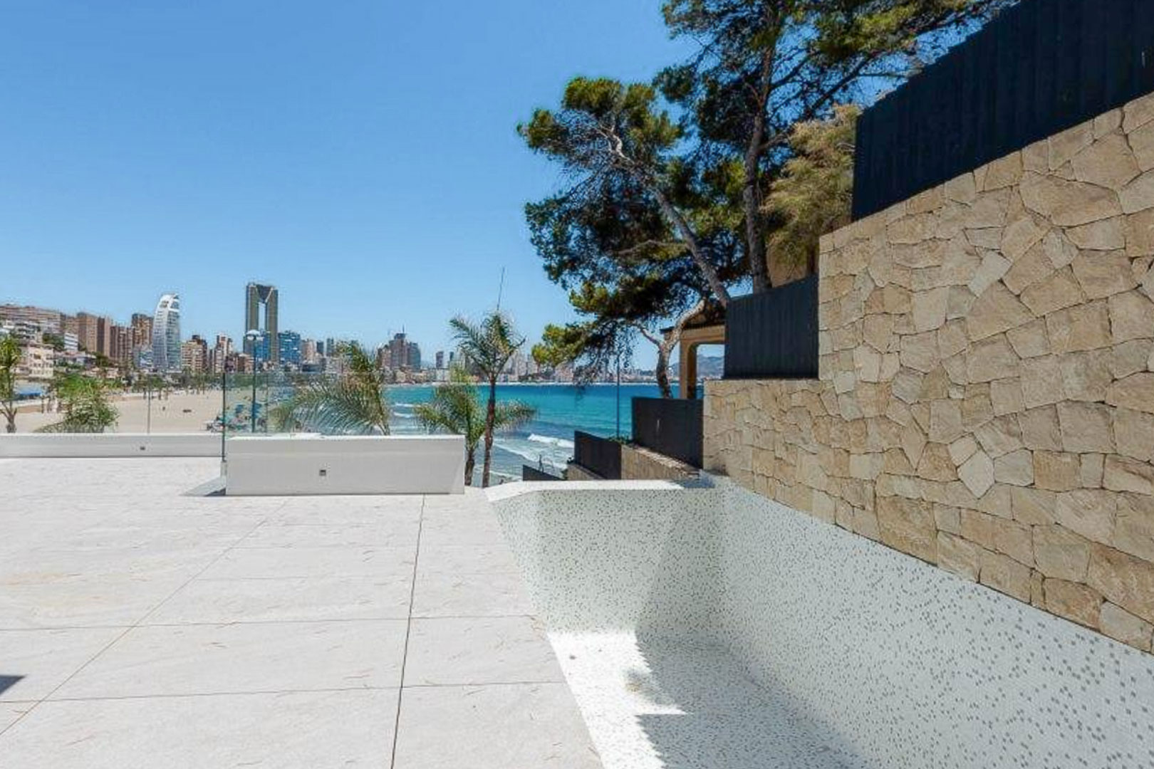 Buitenterras met lichte stenen tegels en een wit mozaïekzwembad, met panoramisch uitzicht op de Middellandse Zee en de skyline van Benidorm.