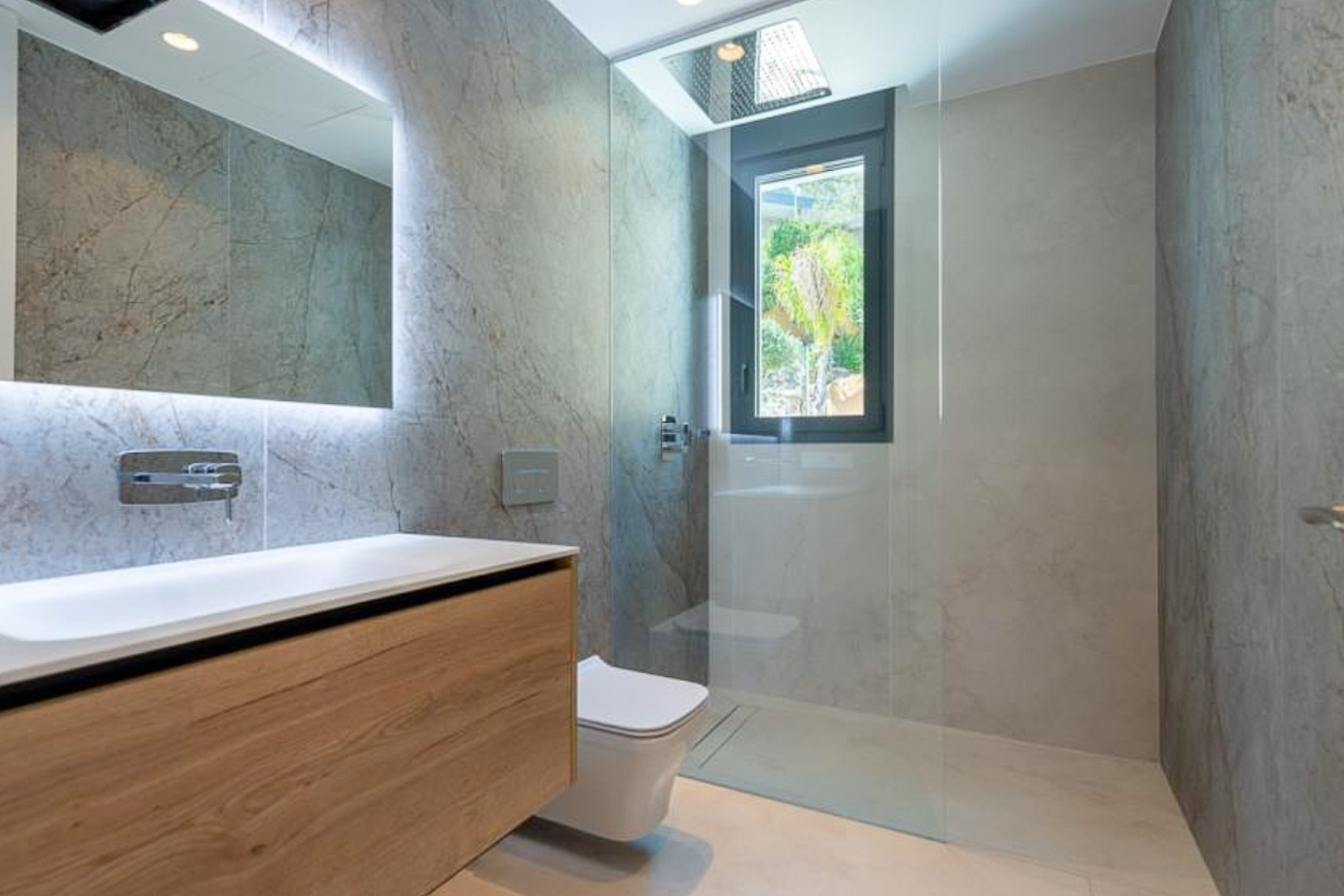 Designbadkamer met wandtegels in natuursteenlook, zwevend houten meubel, verlichte spiegel en inloopdouche met in het plafond geïntegreerde regendouche.