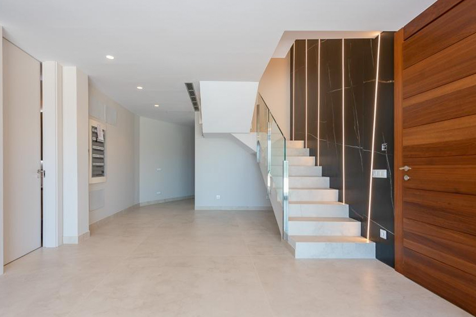 Entreehal met witte stenen trap, glazen balustrade en een donkere marmerlook accentmuur voorzien van verticale LED-verlichtingsstrips.