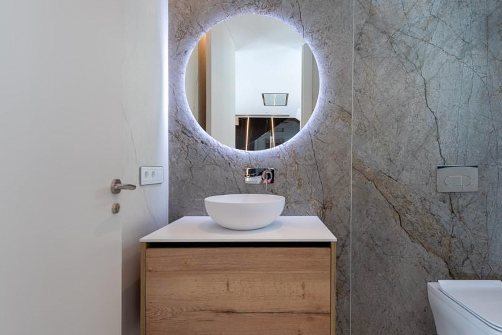 Gastentoilet met grijze marmerlook wandtegels, een ronde verlichte spiegel en een houten wastafelmeubel met opbouwkom en inbouwkraan.