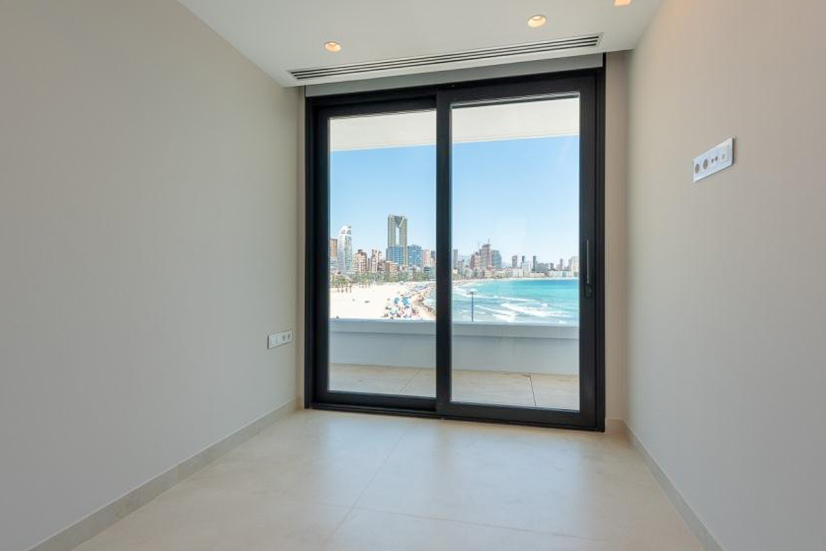 Lichte kamer met een lichte keramische tegelvloer en kamerhoge schuifpui met panoramisch uitzicht op het strand en de skyline van Benidorm.