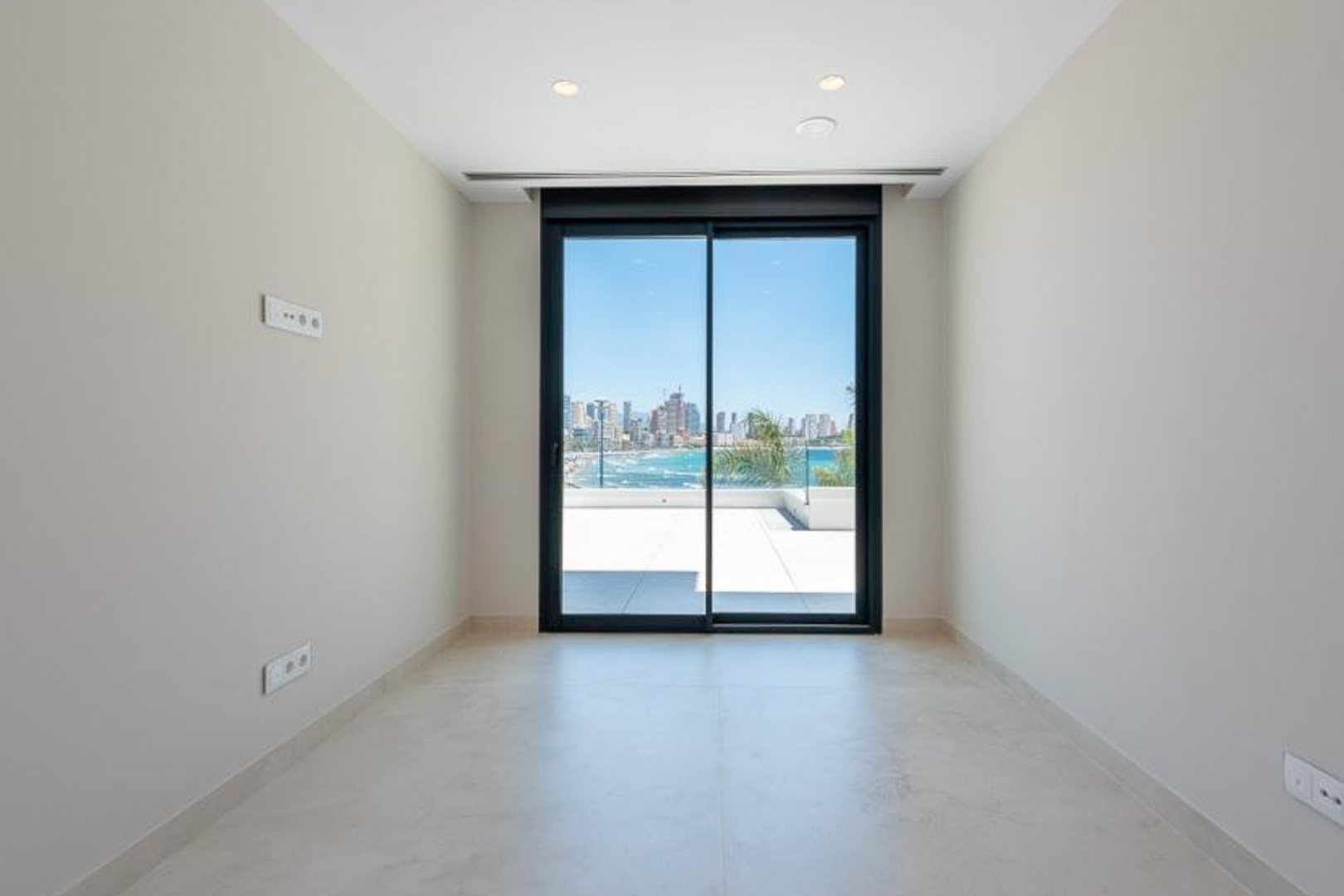 Lichte kamer met porseleinen vloertegels, inbouwspots en een grote glazen schuifdeur die toegang biedt tot het terras met uitzicht op zee en de skyline van Benidorm.