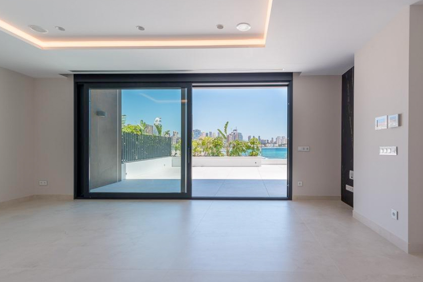 Lichte woonkamer met grootformaat keramische tegels en glazen schuifdeuren die toegang geven tot een privéterras met panoramisch uitzicht op de skyline van Benidorm.