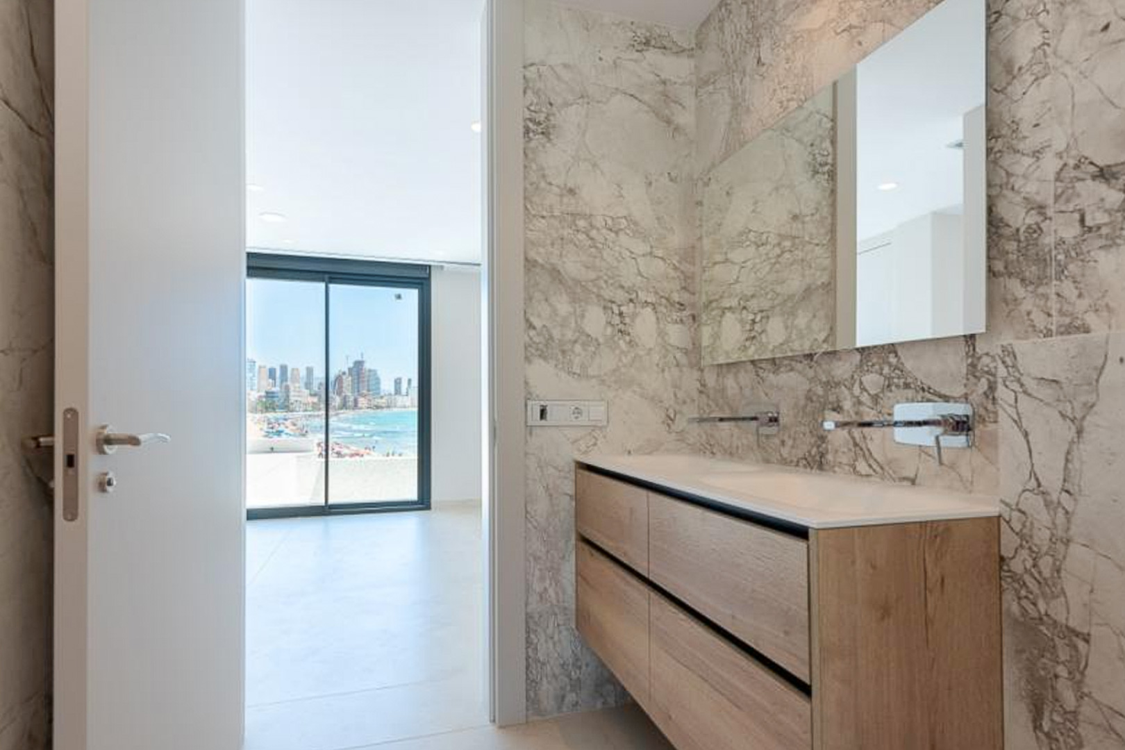 Luxe badkamer met marmeren wanden, een houten dubbel wastafelmeubel en inbouwkranen, met uitzicht op zee via de aangrenzende kamer.