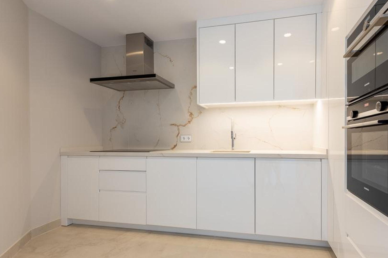 Minimalistische designkeuken met greeploze witte kasten, een marmerlook achterwand met gouden aders en hoogwaardige inbouwapparatuur.
