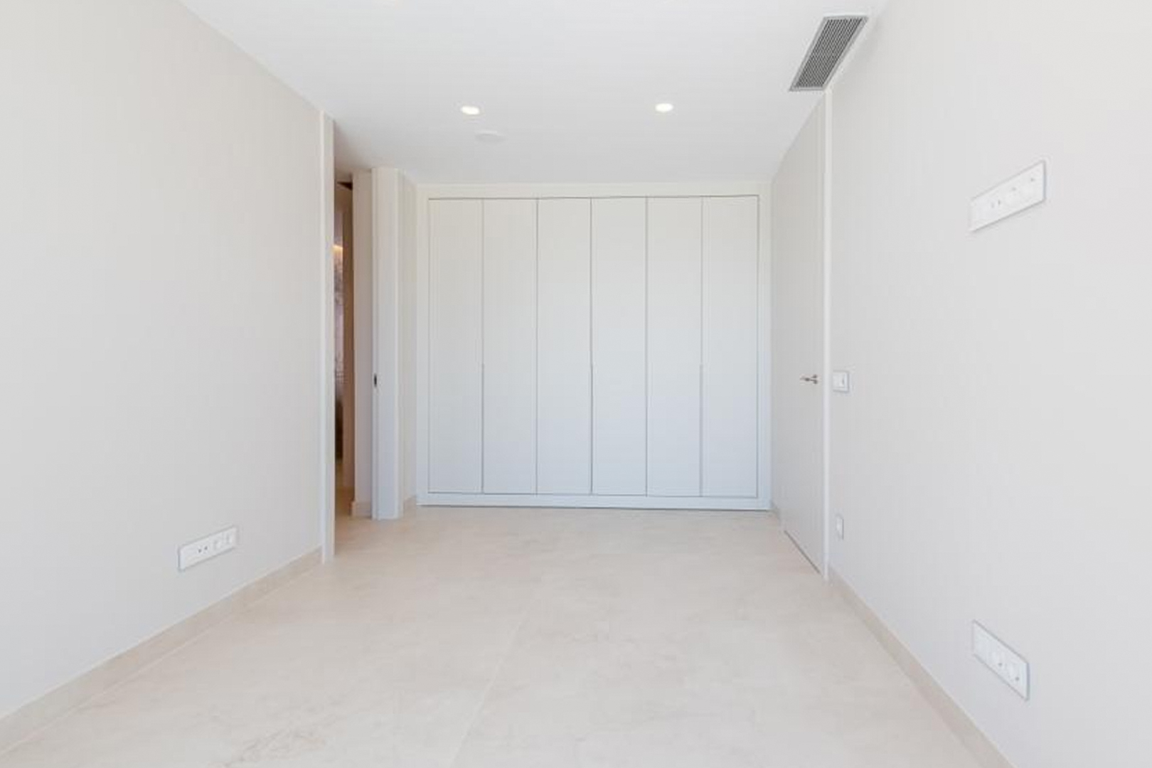 Minimalistische slaapkamer met kamerhoge inbouwkasten, lichte tegelvloer en in het plafond geïntegreerde airconditioning en inbouwspots.