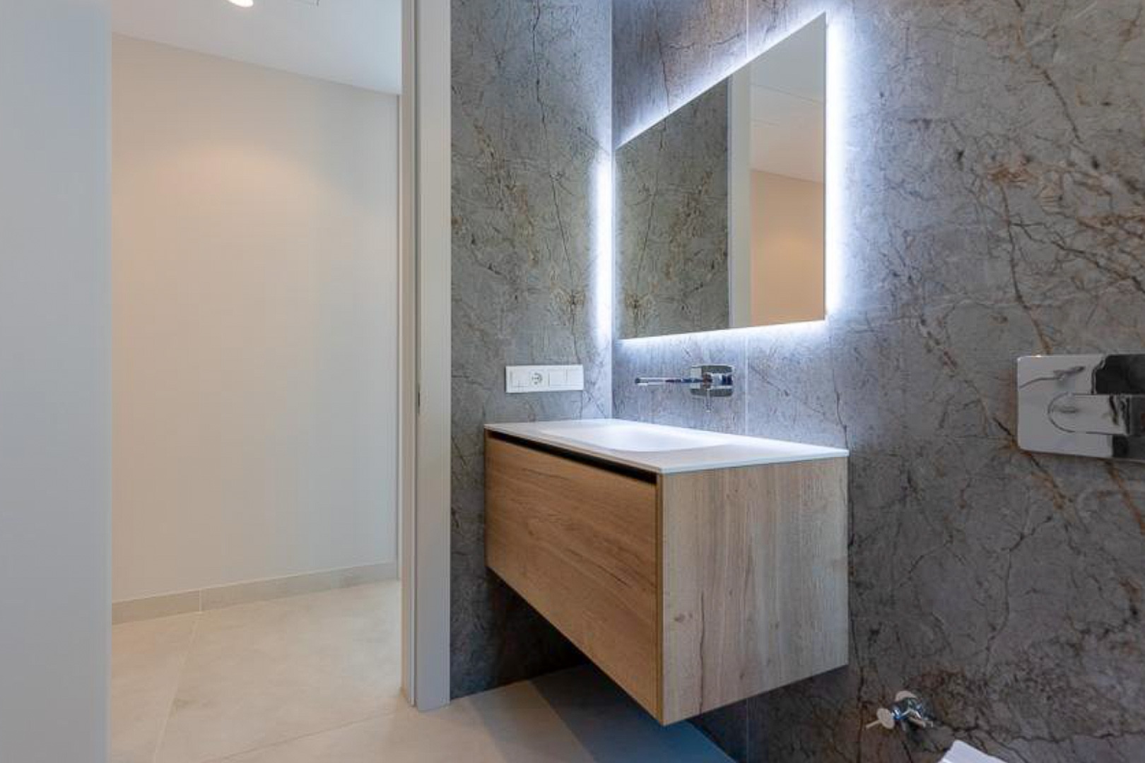Modern gastentoilet met een zwevend houten meubel, geïntegreerde wastafel en verlichte spiegel tegen een grijze wand met marmerlook.