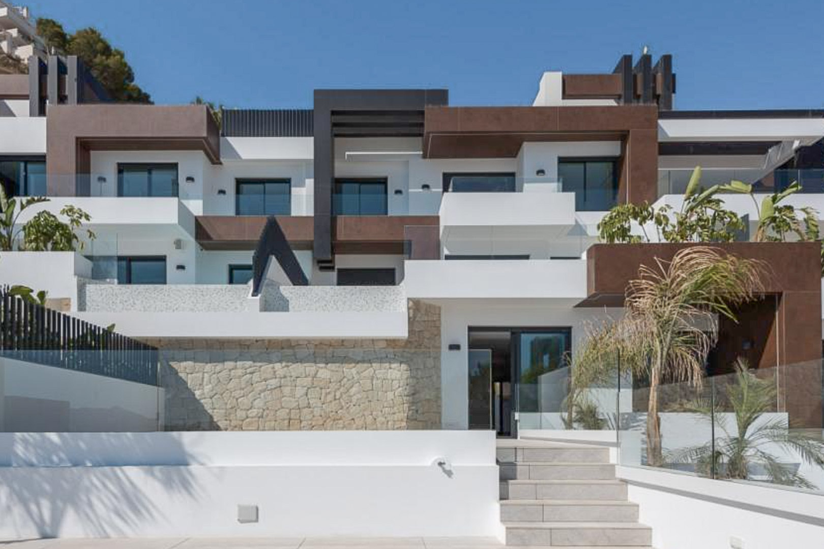 Moderne architecturale gevel met witte en bruine geometrische volumes, natuurstenen muren, glazen balustrades en gelaagde terrassen met palmbomen.