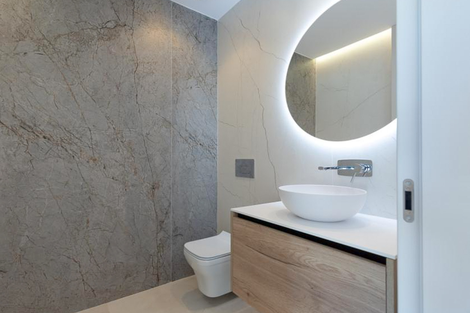Moderne badkamer met zwevend houten meubel, opbouwkom en een ronde verlichte spiegel tegen wanden met marmerlook tegels.