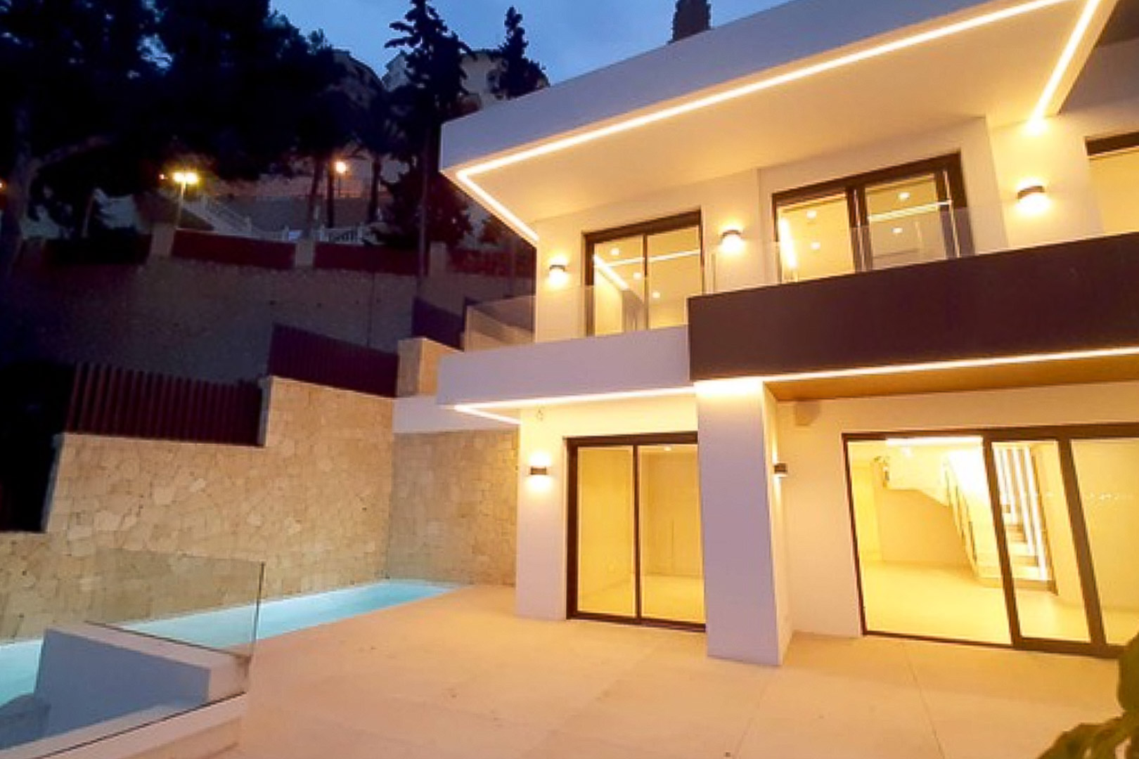 Moderne gevel van twee verdiepingen met geïntegreerde ledverlichting, grote glazen schuifdeuren en een terras met privézwembad naast een natuurstenen muur.