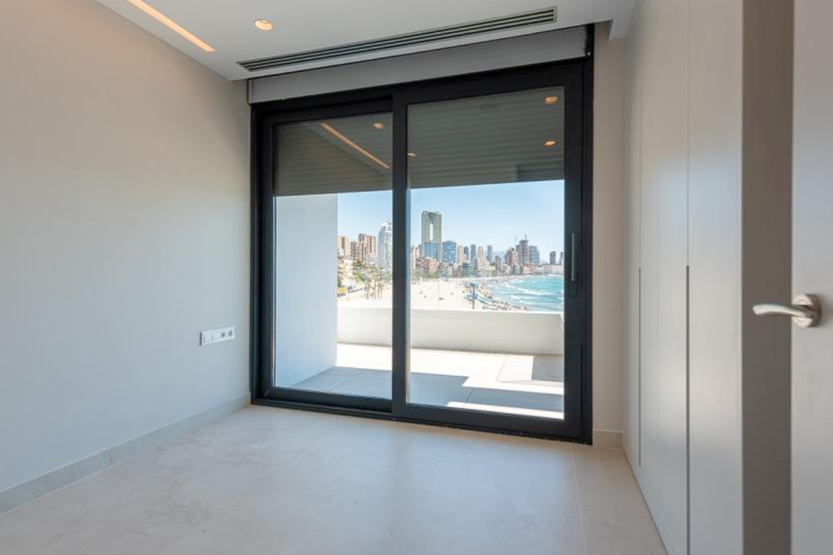 Slaapkamer met kamerhoge inbouwkasten en een schuifpui die toegang biedt tot het terras met uitzicht op het strand en de skyline van Benidorm.