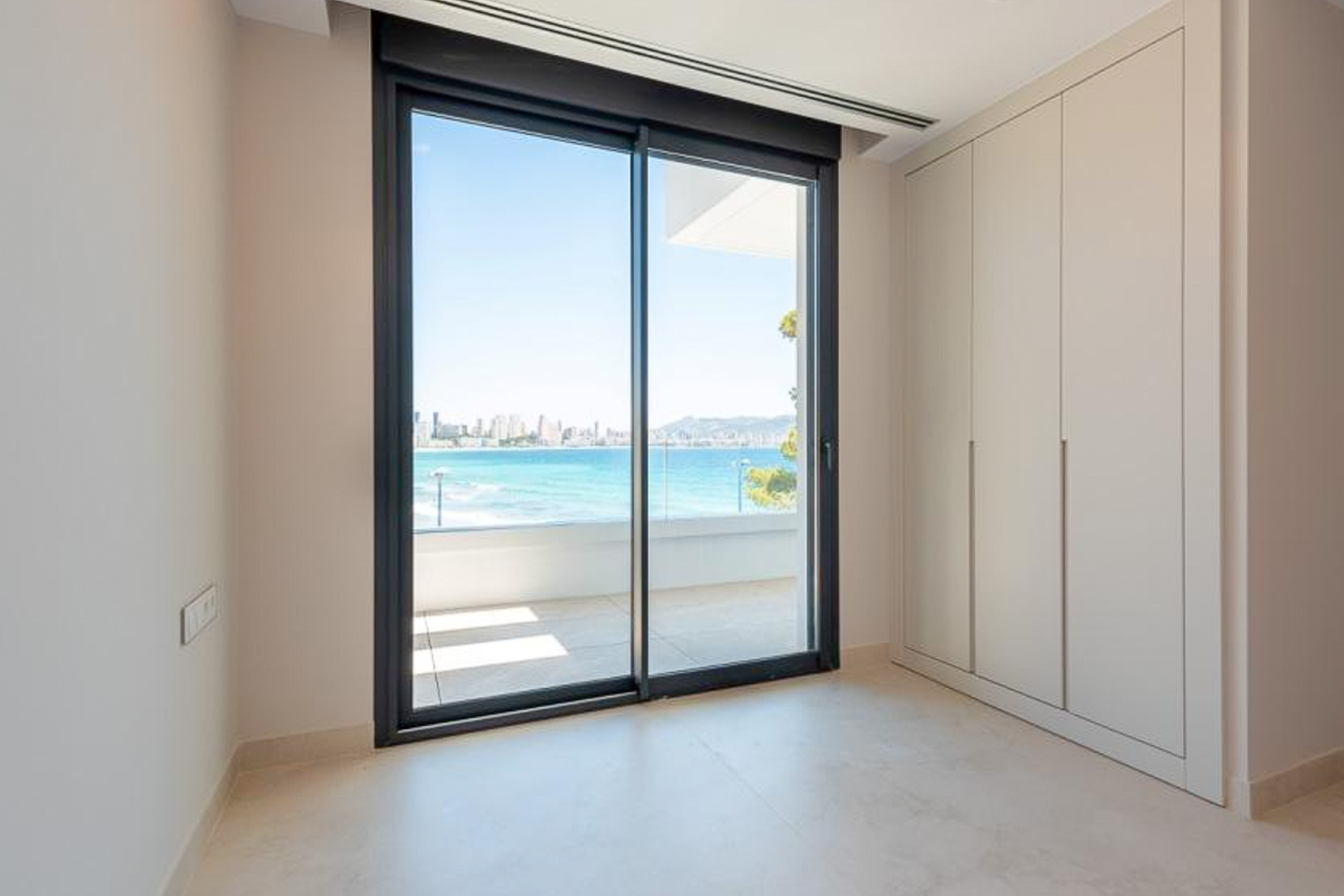 Slaapkamer met kamerhoge inbouwkasten en grote schuifpui naar een terras met uitzicht op de Middellandse Zee en de skyline van Benidorm.