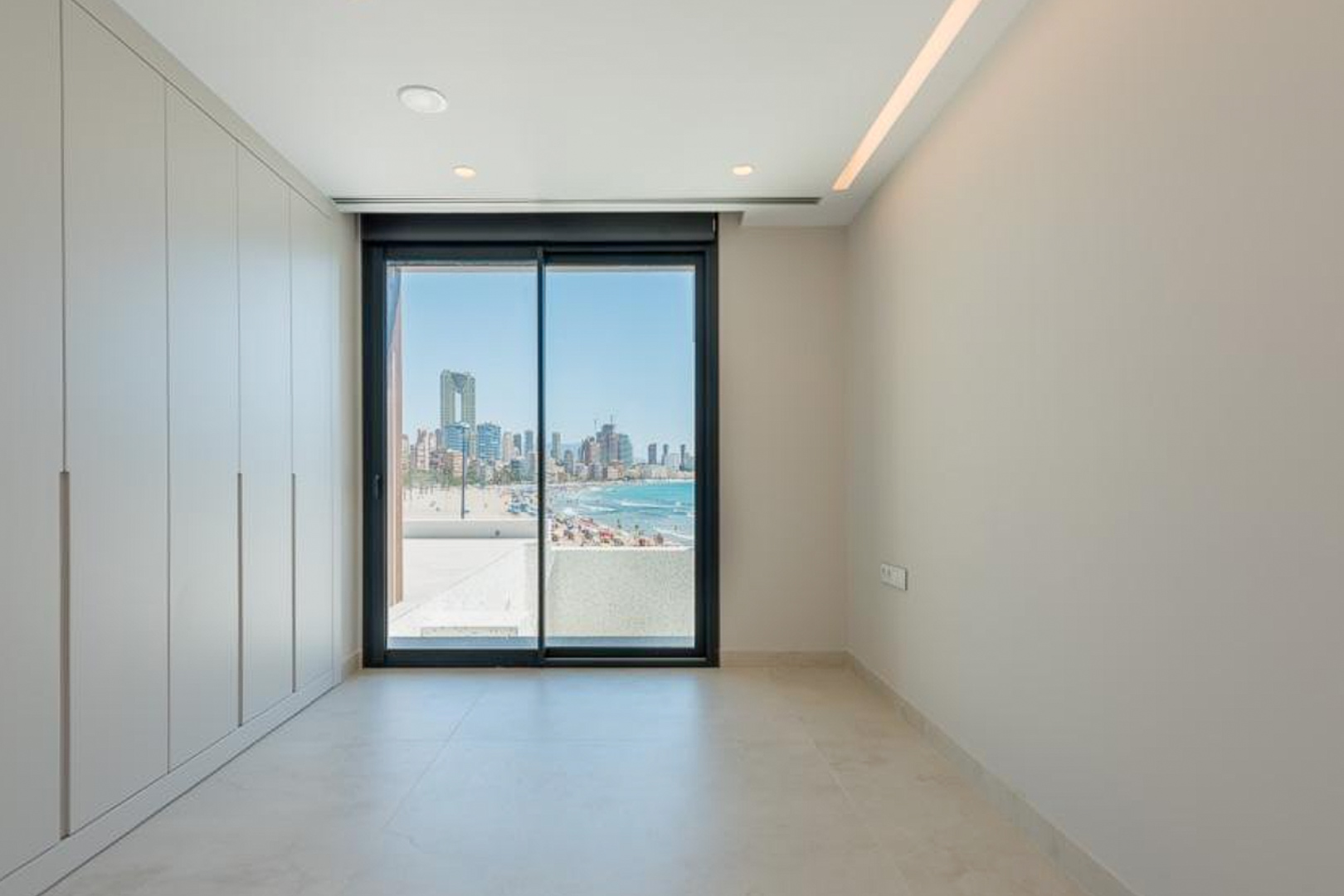Slaapkamer met kamerhoge inbouwkasten, geïntegreerde ledverlichting en uitzicht op het Poniente-strand en de skyline van Benidorm.