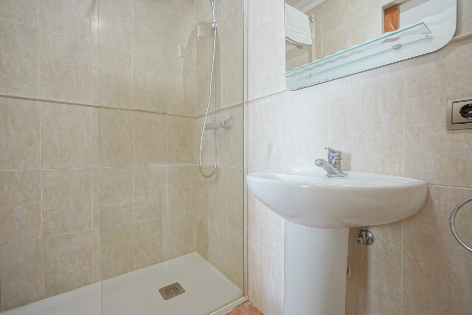 Badkamer met beige wandtegels, voorzien van een witte kolomwastafel en een inloopdouche met glazen wand en chromen kranen.