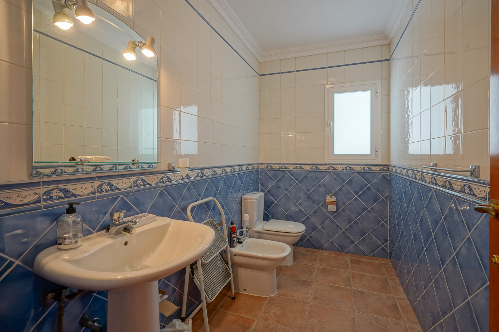 Badkamer met blauw-witte wandtegels, een terracotta vloer en klassiek sanitair inclusief een kolomwastafel, bidet en toilet met geïntegreerde spiegelverlichting.