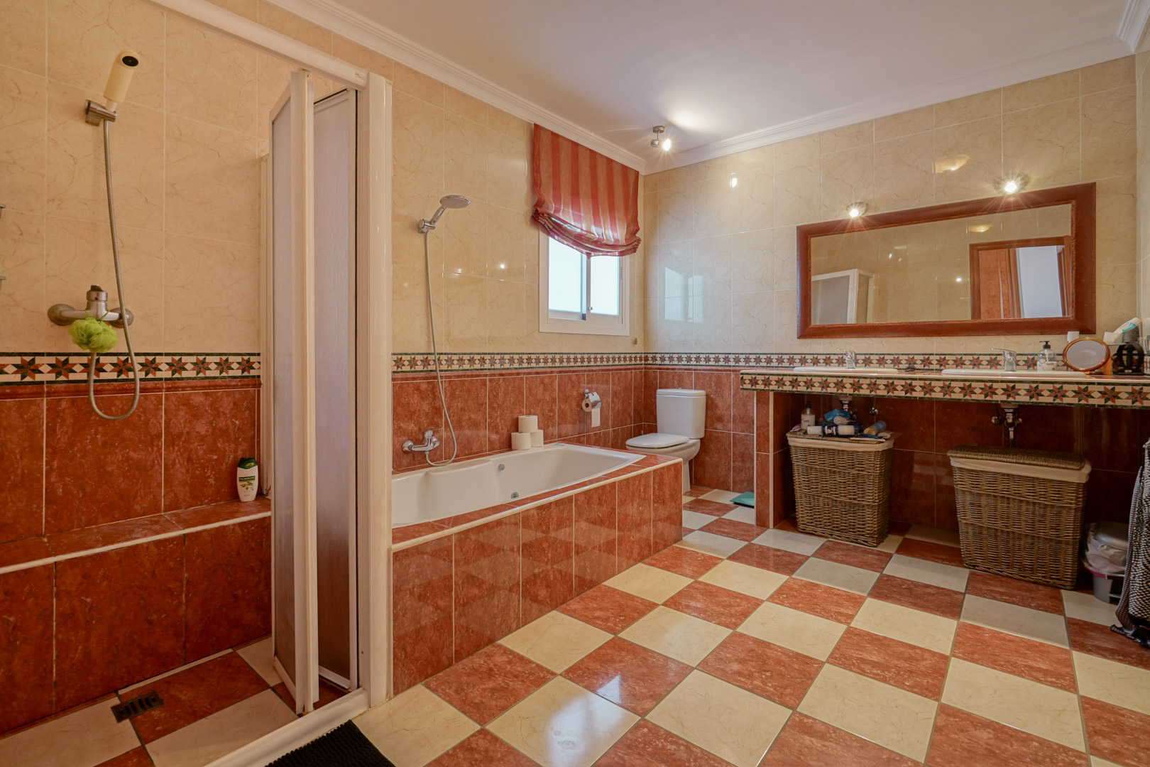 Badkamer met crème en roodachtige tegels, voorzien van een inbouwbad, aparte douche en een dubbele wastafel met een grote spiegel met houten lijst.