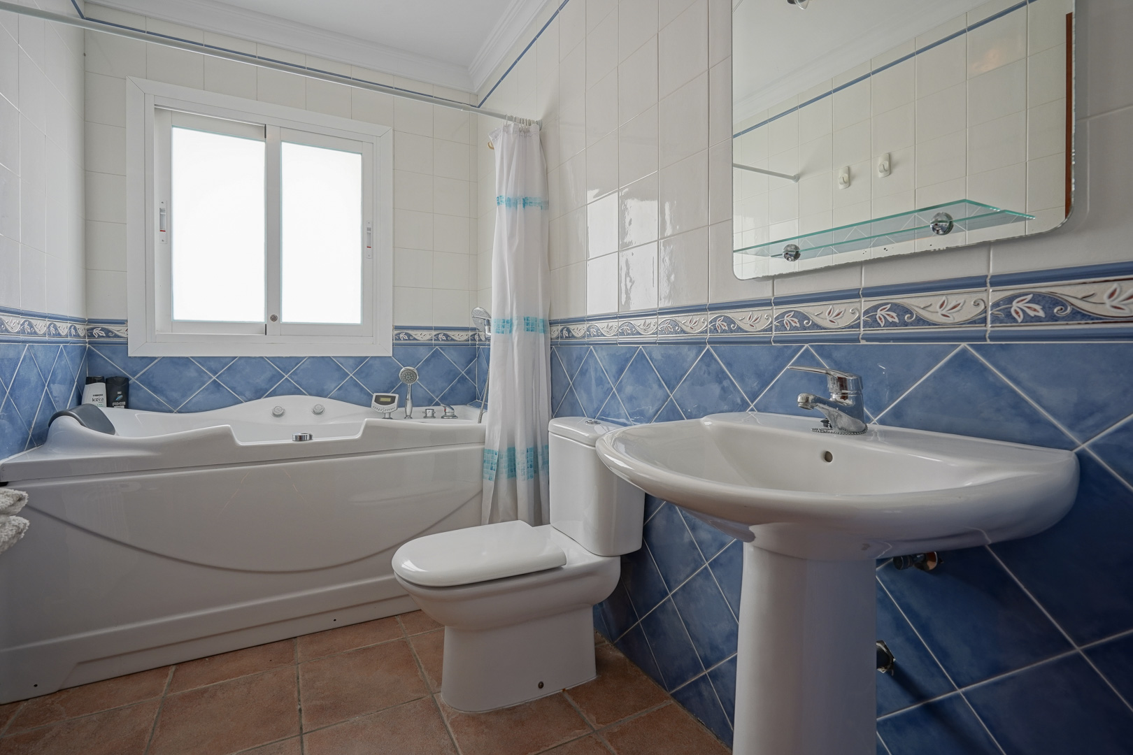 Badkamer met whirlpool ligbad, kolomwastafel en blauw-witte wandtegels. Voorzien van een terracotta vloer en een raam voor natuurlijk lichtinval.