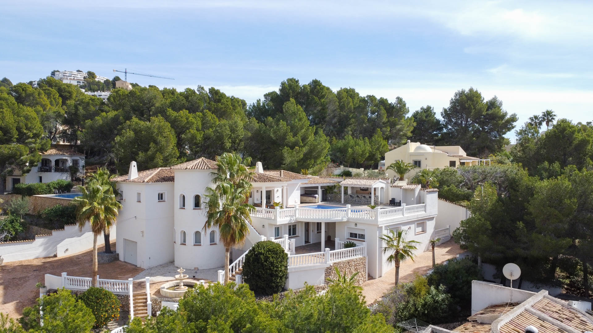 Externe weergave van de mediterrane villa met witte gevel, ronde toren en terrassen met balustrades, omgeven door pijnbomen en een verhoogd zwembad.