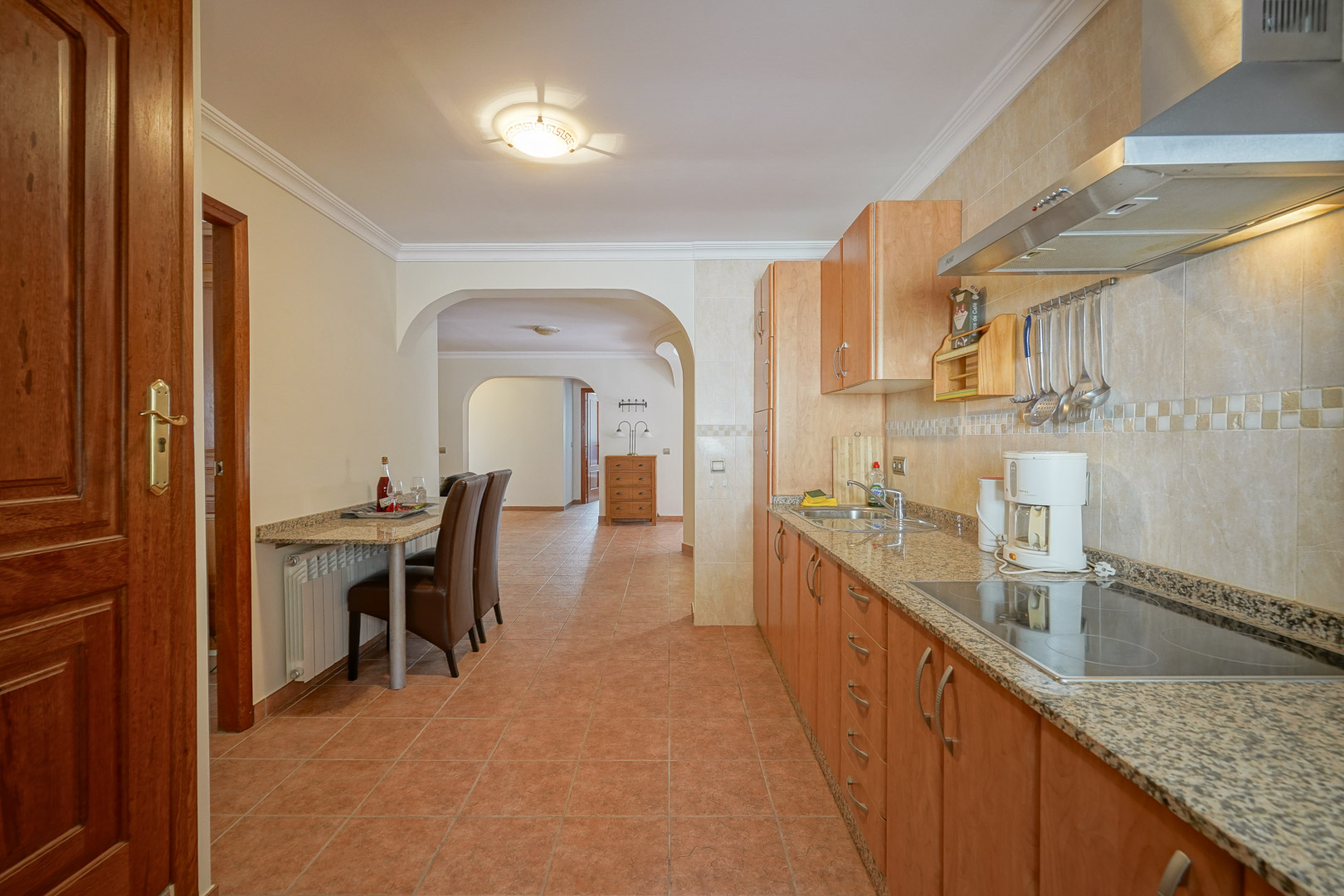 Functionele keuken met houten kasten, granieten werkbladen en een keramische kookplaat. Voorzien van een geïntegreerde ontbijtbar en tegelvloeren die via bogen aansluiten op de woning.