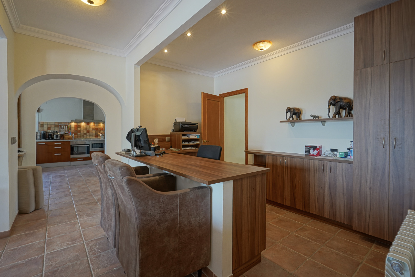 Kantoorruimte met op maat gemaakte houten kasten, terracotta vloertegels en karakteristieke bogen die doorgang bieden naar de keuken.