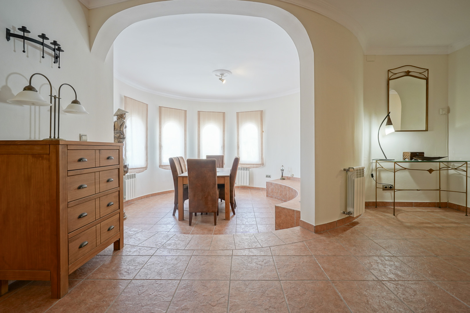 Lichte eethoek met boogvensters en terracotta vloertegels. De ronde architecturale vormgeving is voorzien van houten meubilair en sluit direct aan op de hal.