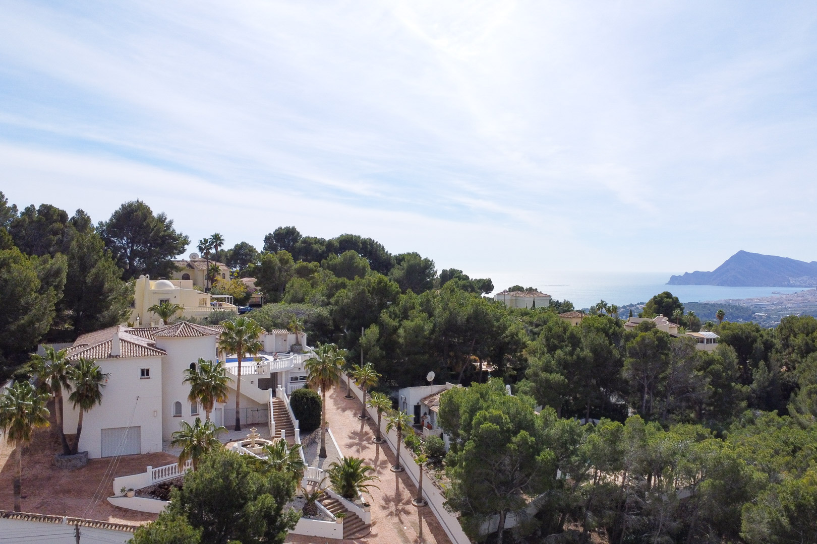 Luchtfoto van de witte mediterrane villa met ronde toren, omgeven door pijnbomen en palmen, met een panoramisch uitzicht over de kustlijn en de zee.