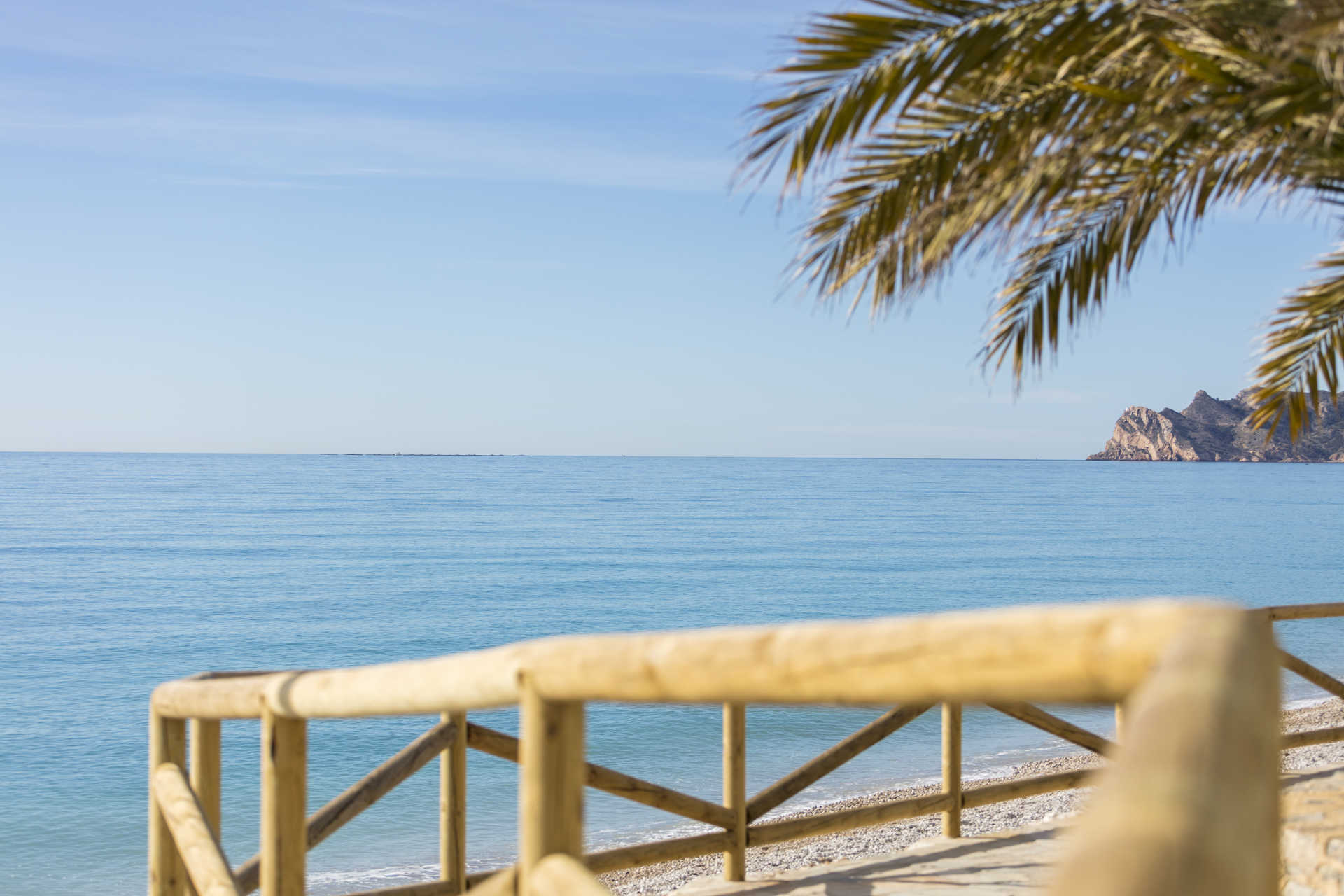 Panoramisch uitzicht op de Middellandse Zee vanaf de kust van Altea, met houten leuningen, een kiezelstrand en rotsachtige kliffen in de verte.