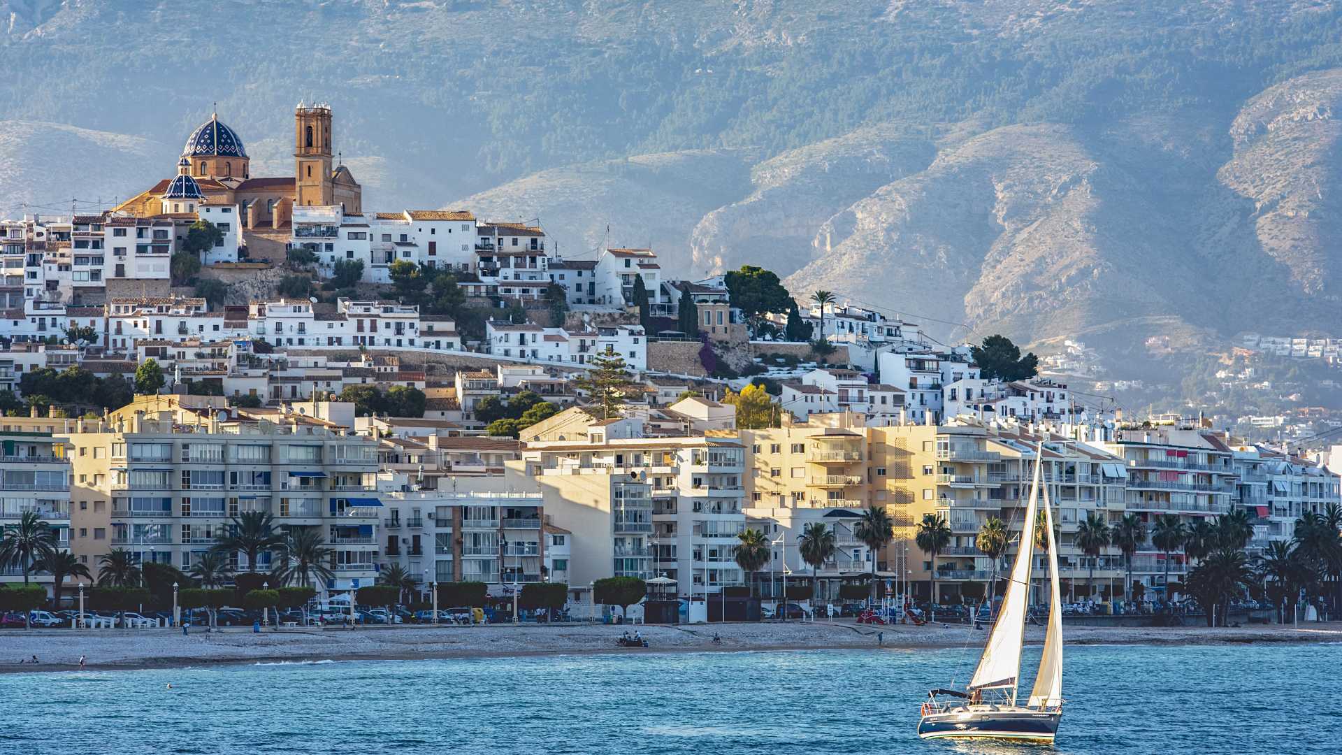 Panoramisch uitzicht op de oude stad van Altea met de iconische blauwe koepelkerk, de Sierra de Bernia op de achtergrond en een zeilboot op de Middellandse Zee.