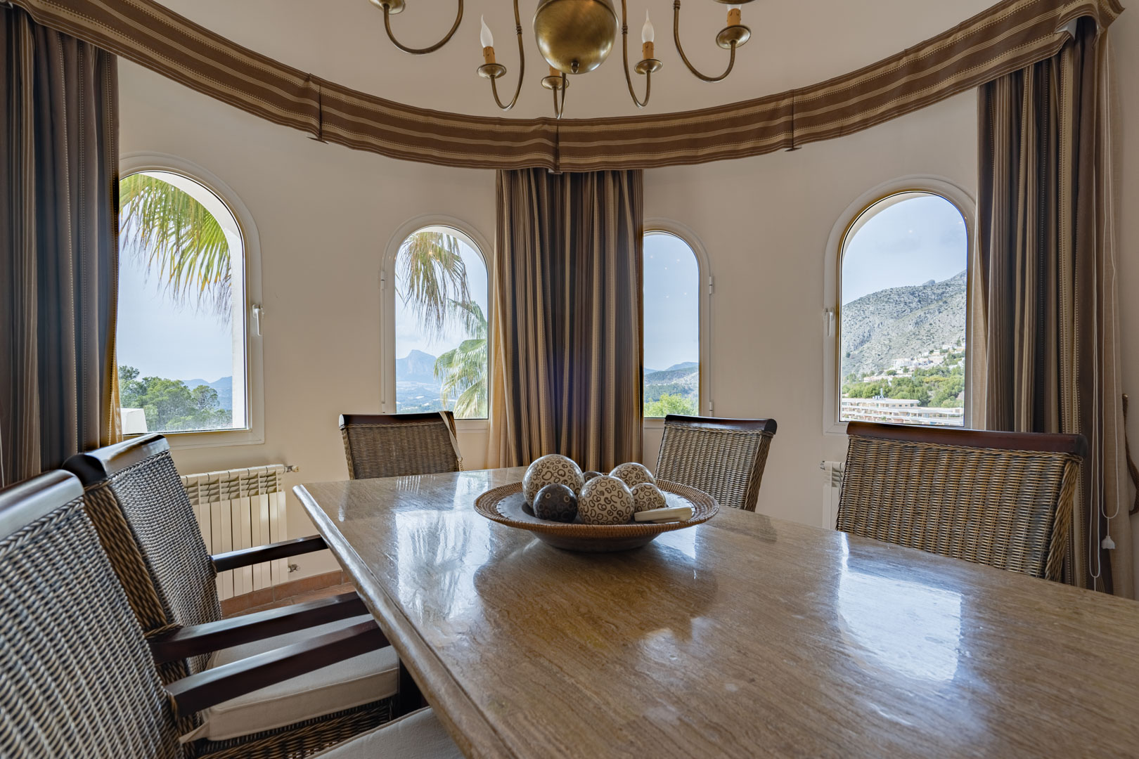 Ronde eetkamer met een gepolijste stenen tafel en rotan stoelen. Vier boogvensters bieden een panoramisch uitzicht op de Sierra de Bernia en het berglandschap van Altea.