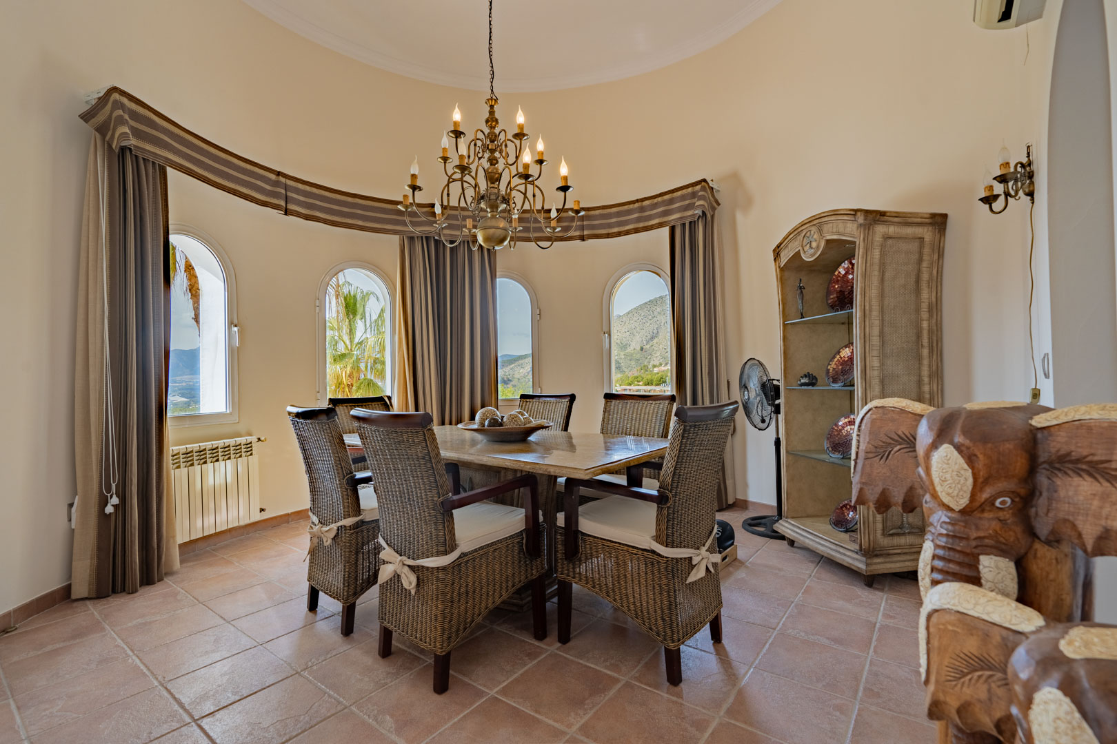 Ronde eetkamer met een stenen tafel, rotan stoelen en een klassieke kroonluchter. Vier boogvensters bieden uitzicht op het omliggende Sierra de Bernia-gebergte.