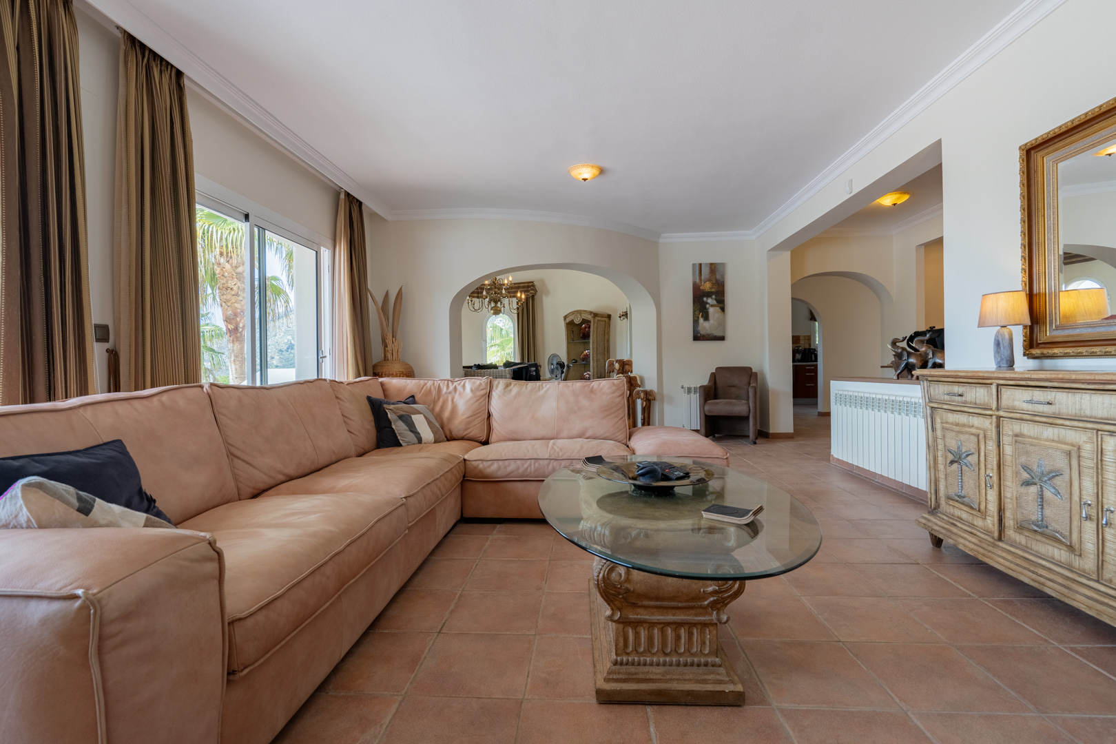 Ruime woonkamer met terracotta vloertegels, een grote lederen hoekbank en karakteristieke bogen. De glazen salontafel en het houten dressoir passen bij de mediterrane stijl.