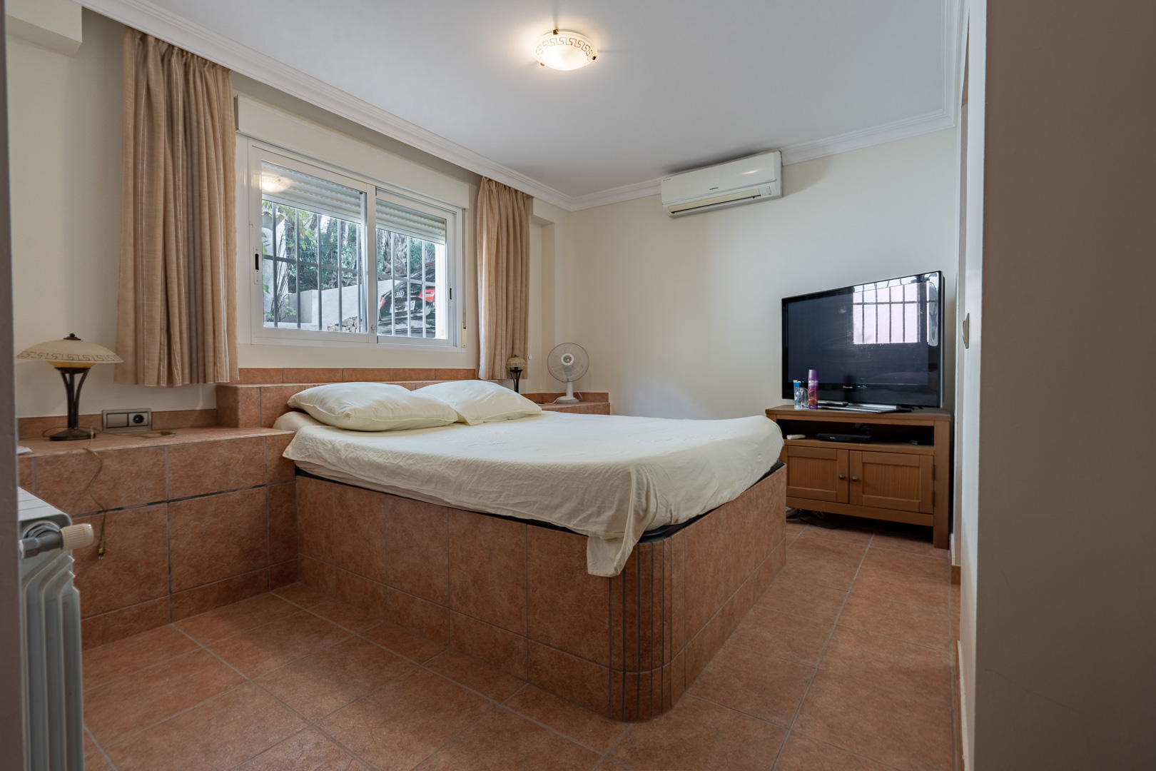 Slaapkamer met een betegeld bedframe en bijpassende vloer, voorzien van airconditioning, radiatorverwarming en een houten tv-meubel.