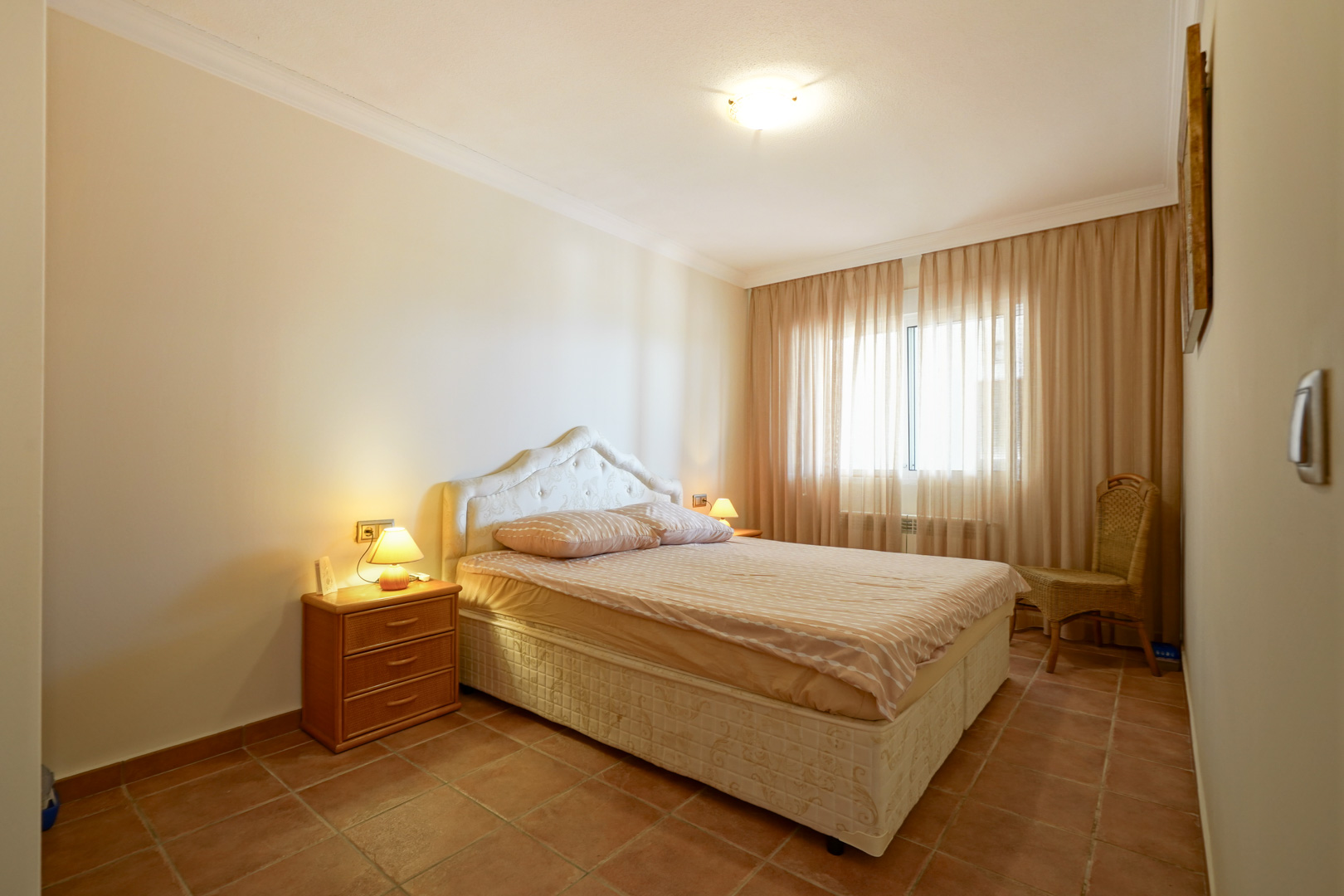 Slaapkamer met terracotta tegelvloer, tweepersoonsbed met gecapitonneerd hoofdbord, houten nachtkastje en veel lichtinval via de grote raampartij.