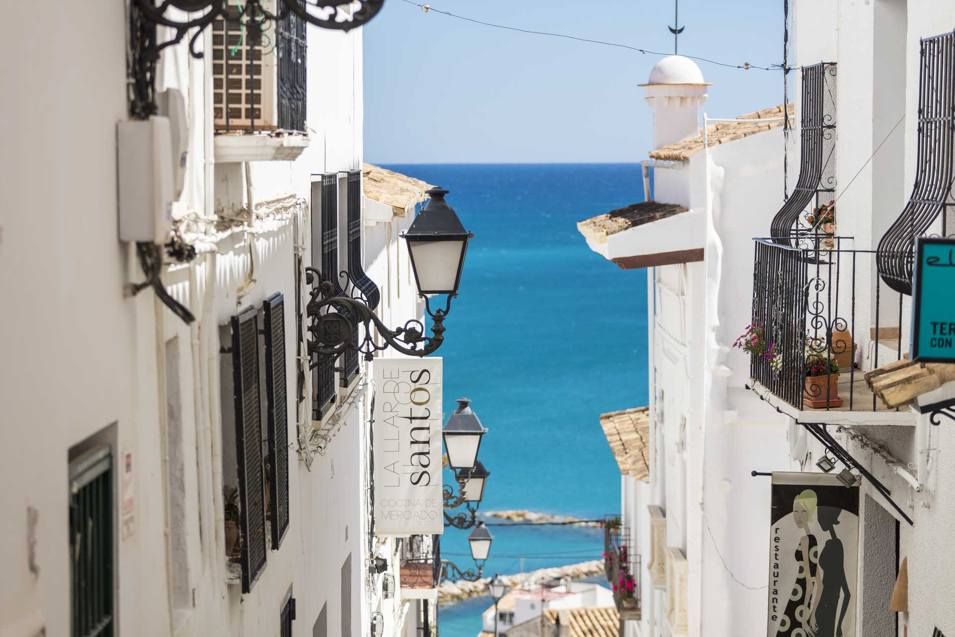 Uitzicht op een smalle straat in de oude stad van Altea, met witgekalkte gevels, smeedijzeren lantaarns en een doorkijk naar de azuurblauwe Middellandse Zee.