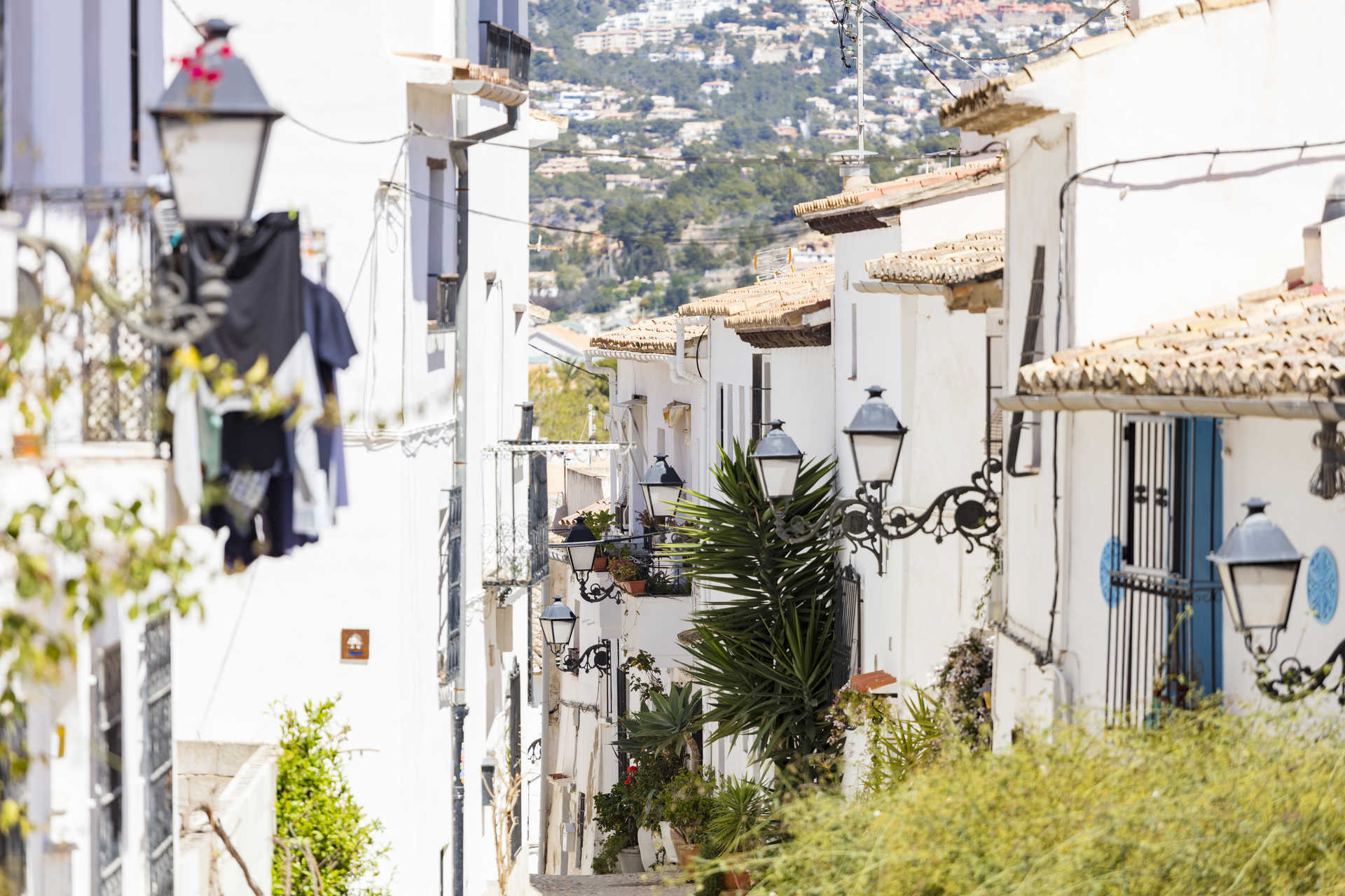 Zicht op een smalle straat in de oude stad van Altea, met traditionele witgekalkte huizen, smeedijzeren lantaarns en uitzicht op de omliggende heuvels.