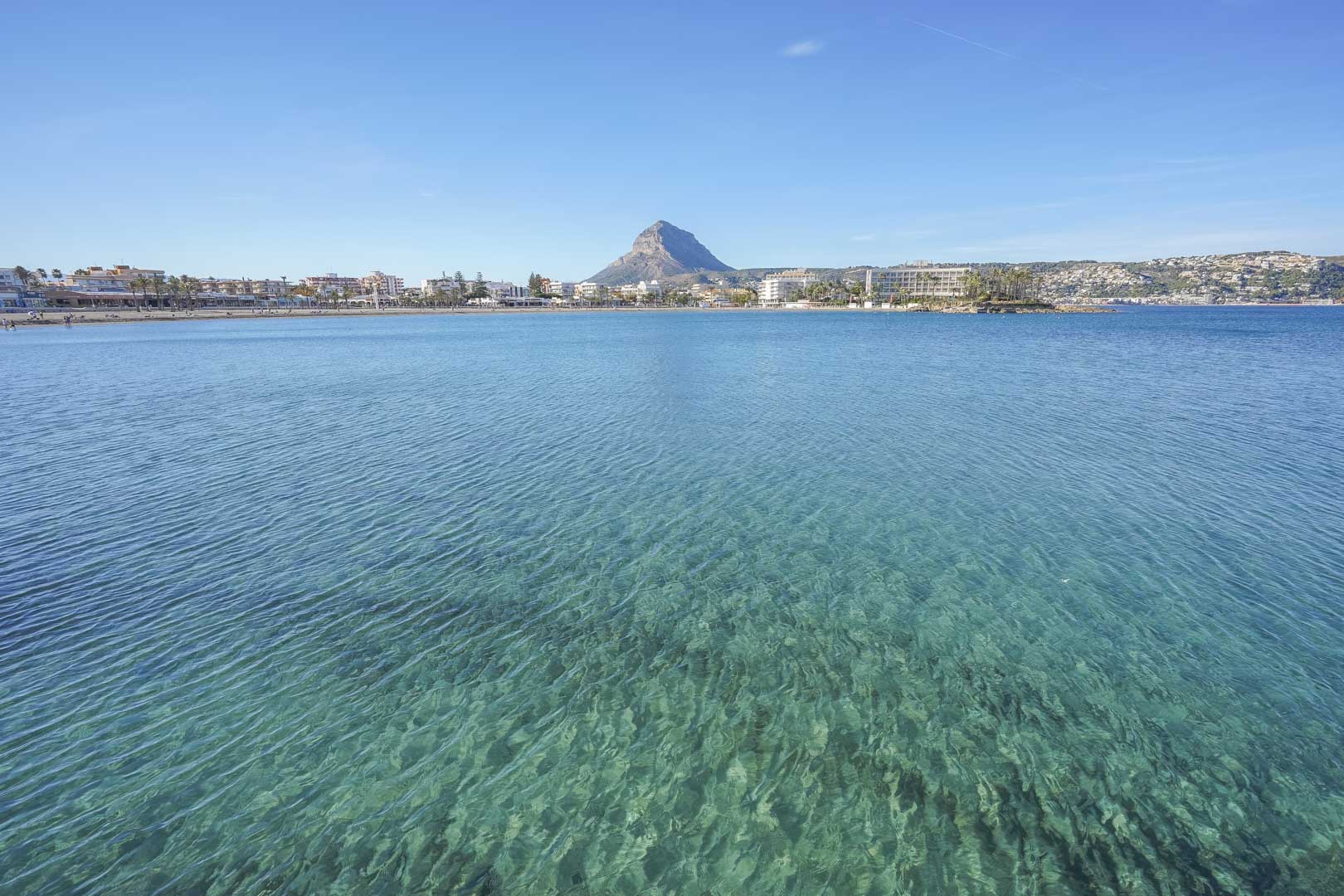 Panoramisch uitzicht op de baai van Jávea met kristalhelder water, de kustlijn en de karakteristieke Montgó-berg op de achtergrond.