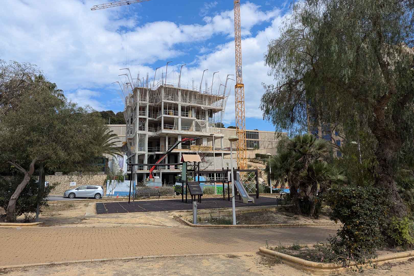 Huidige voortgang van de bouw in Calpe, met de gewapende betonstructuur en bouwkranen gelegen naast een openbaar park met speeltoestellen.