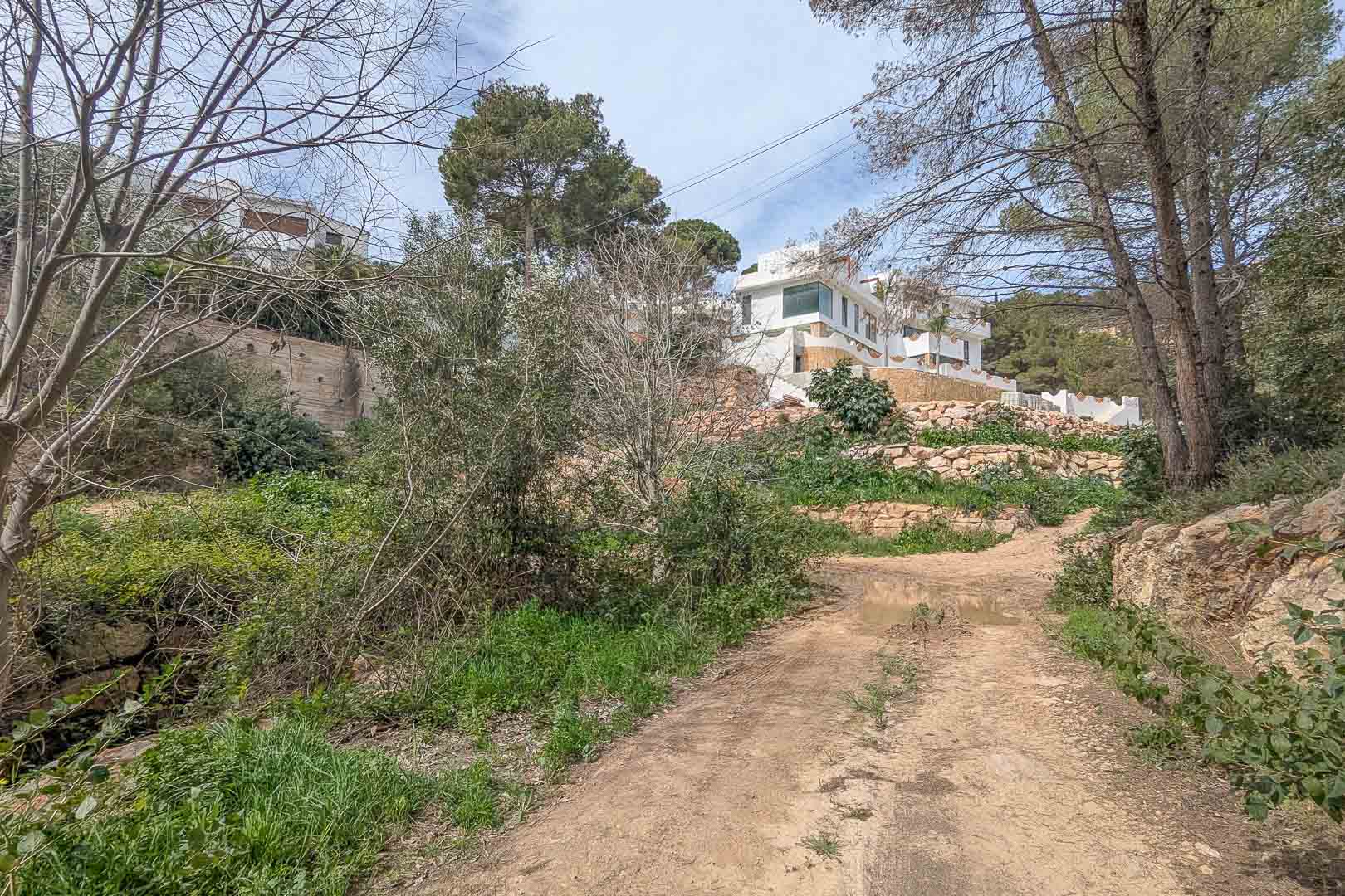 Buitenaanzicht vanaf het toegangspad met de verhoogde kavel, natuurstenen muren en de witte villa gelegen tussen de karakteristieke pijnbomen.
