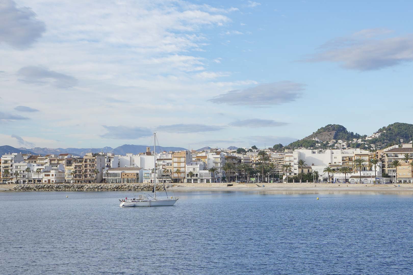 Panoramisch uitzicht op de haven van Jávea met een zeilboot in de baai, de boulevard met witte bebouwing en de bergen op de achtergrond.