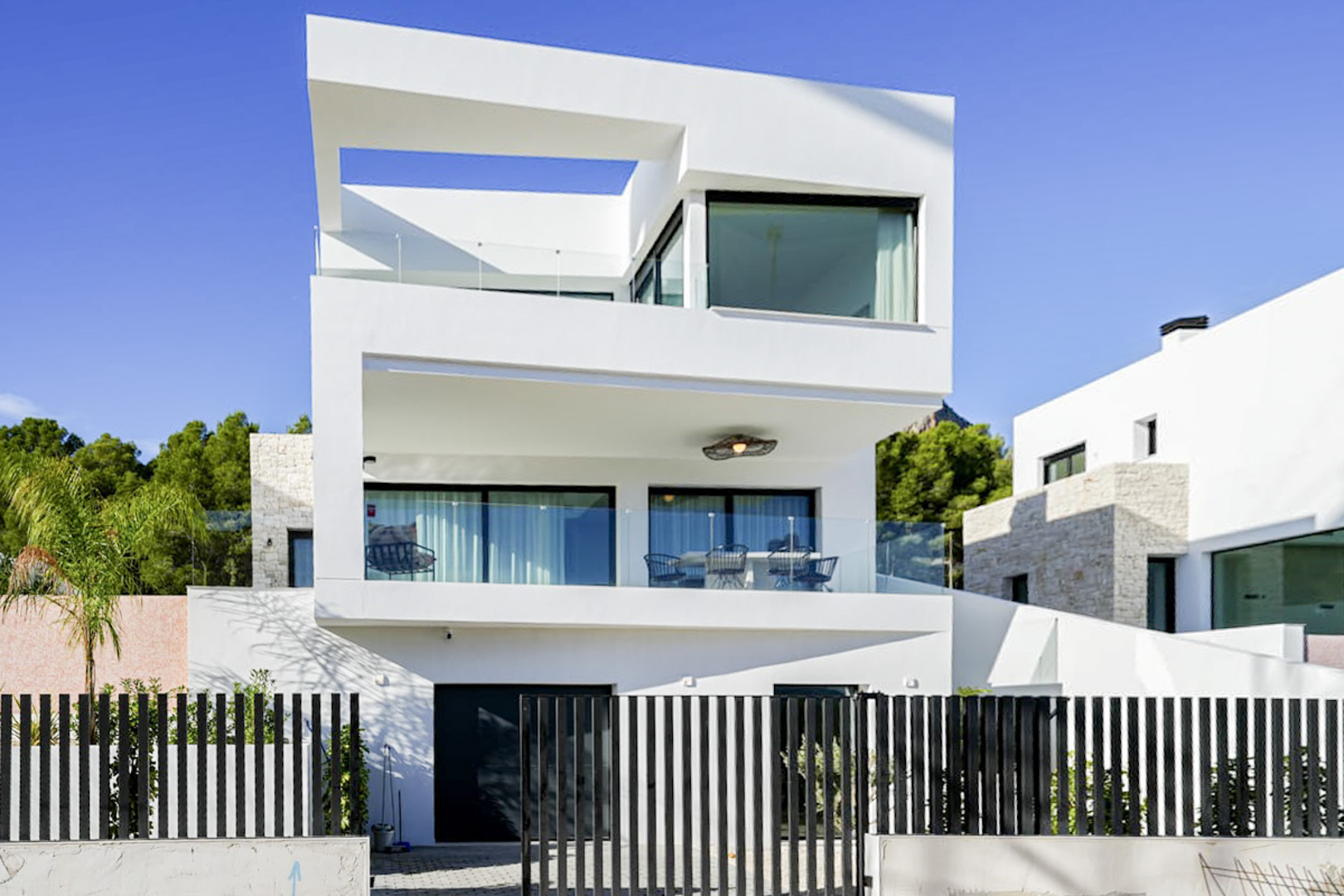 Exterieur van een moderne villa met drie verdiepingen, balkons, glazen balustrades en een zwart lattenhek.