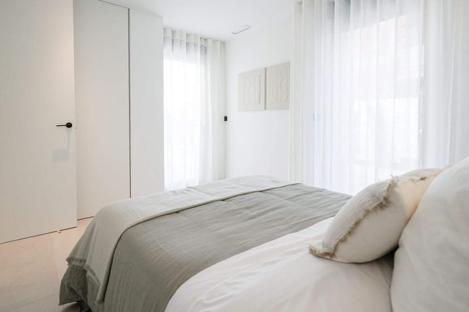 Lichte slaapkamer met grijs en wit beddengoed, transparante gordijnen en minimalistische wanddecoratie.
