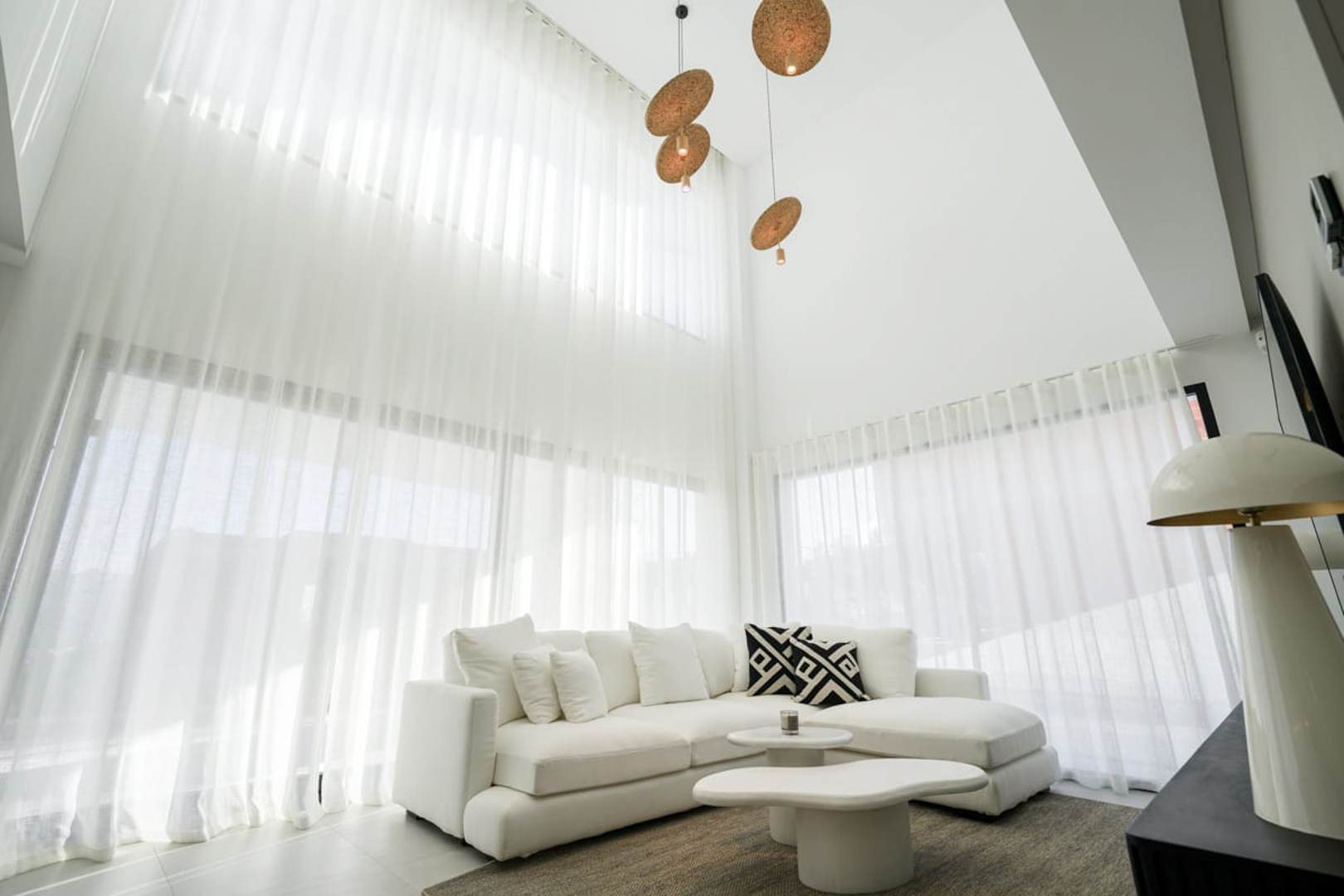 Lichte woonkamer met witte gordijnen, hoekbank en design hanglampen, waardoor een moderne, minimalistische sfeer ontstaat.