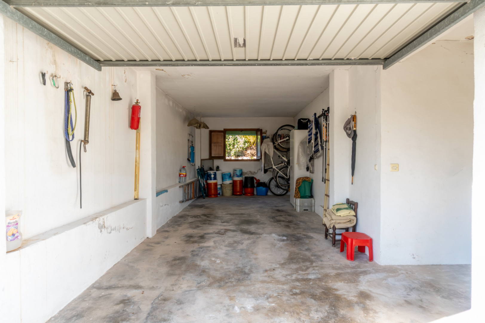 Ruime afgesloten garage met betonnen vloer en witte muren, die veel opbergruimte en natuurlijk licht biedt via een raam aan de achterzijde.