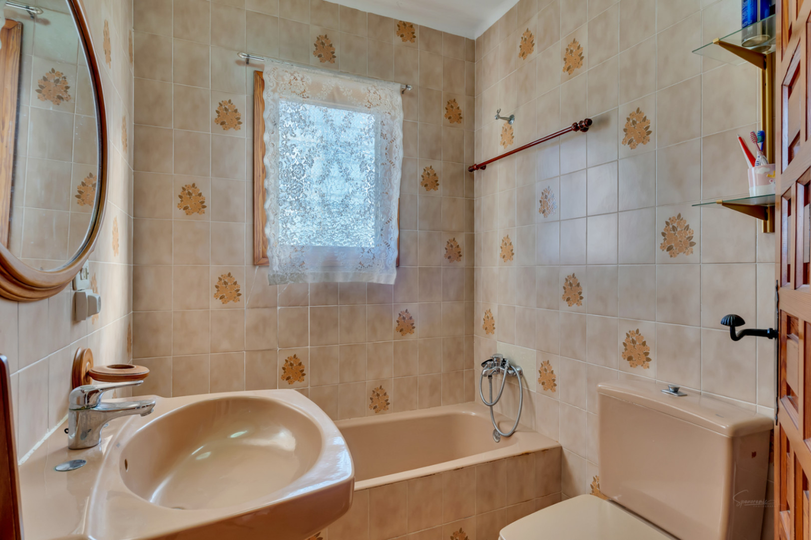 Traditionele badkamer met beige bloemenpatroon tegels, bijpassend sanitair en een ingebouwd bad. Voorzien van een ovale spiegel met houten lijst en natuurlijk licht.