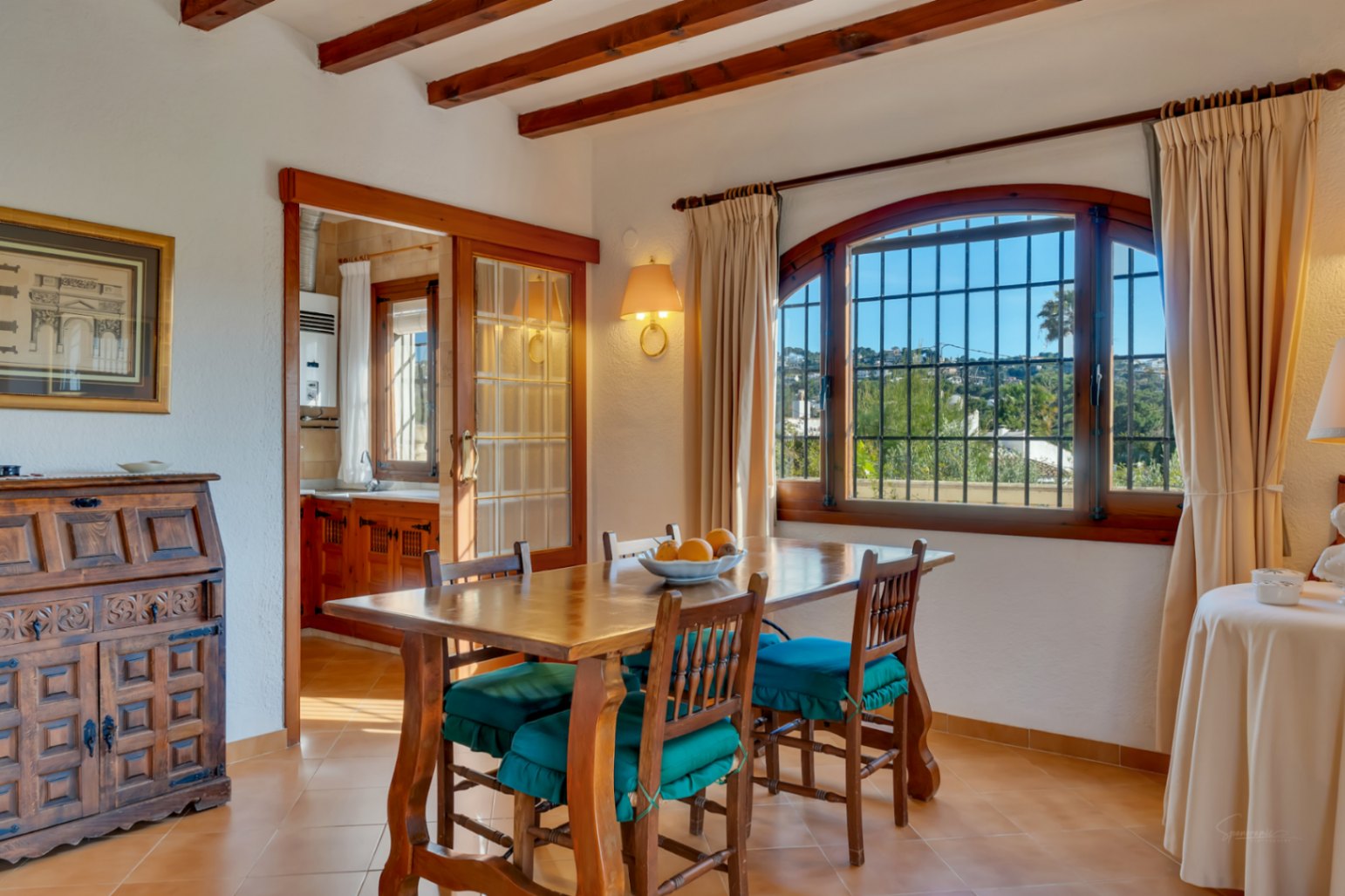 Traditionele eetkamer met houten balkenplafond, massief houten tafel en een boogvenster. Een glazen schuifdeur biedt toegang tot de naastgelegen keuken.