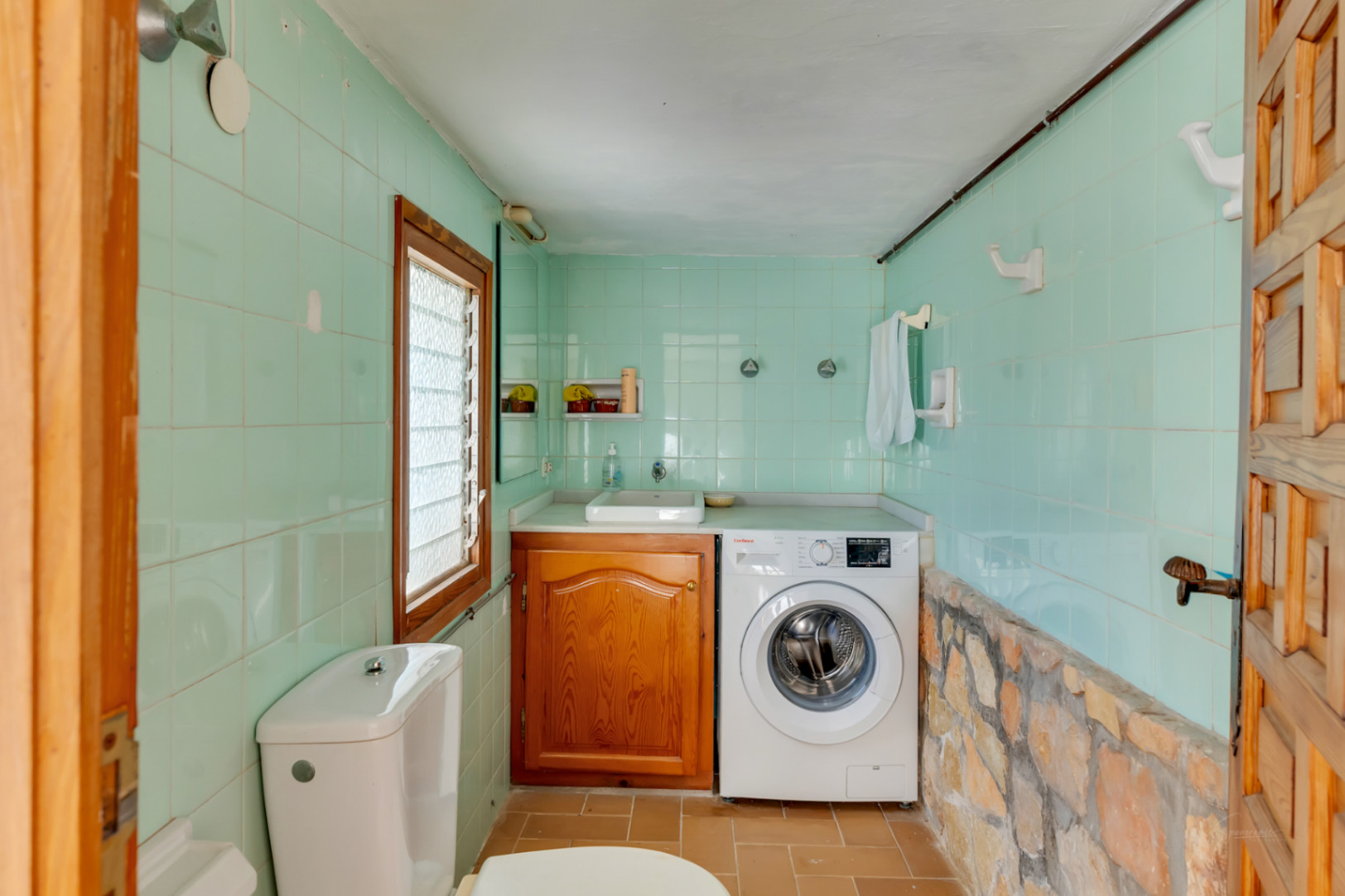 Wasruimte en toilet met mintgroene tegels, een natuurstenen wand, houten kastje en een geïntegreerde wasmachine.