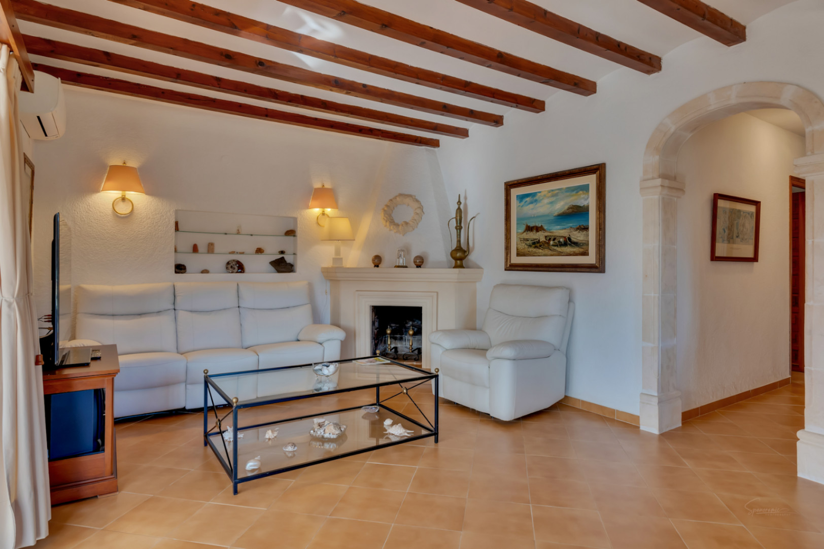 Woonkamer met houten plafondbalken, een klassieke open haard en een boog van tosca-natuursteen. Witte meubels op een traditionele tegelvloer zorgen voor een lichte sfeer.