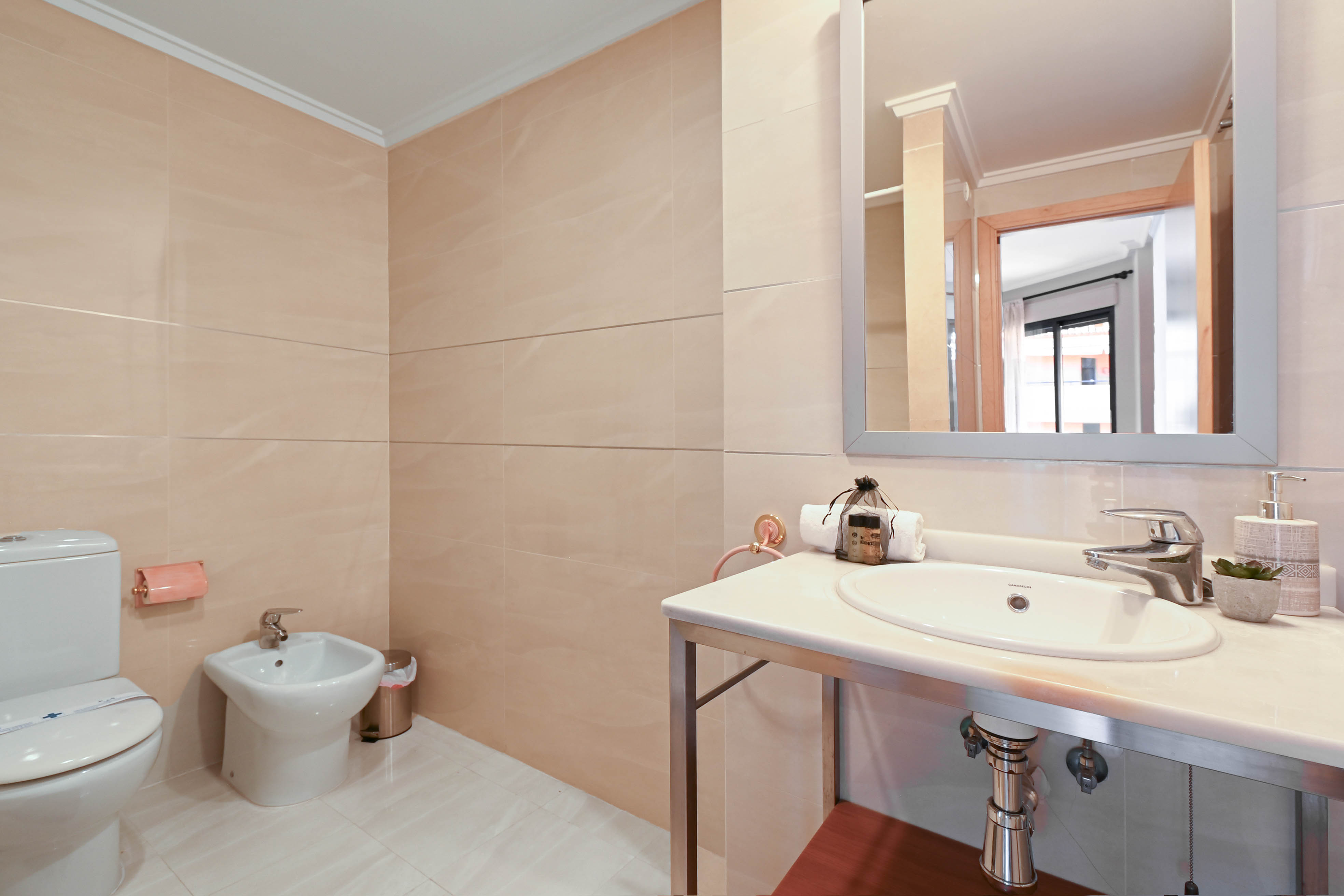 Badkamer met beige wandtegels, voorzien van een bidet, toilet en een witstenen wastafelmeubel op een chromen onderstel.