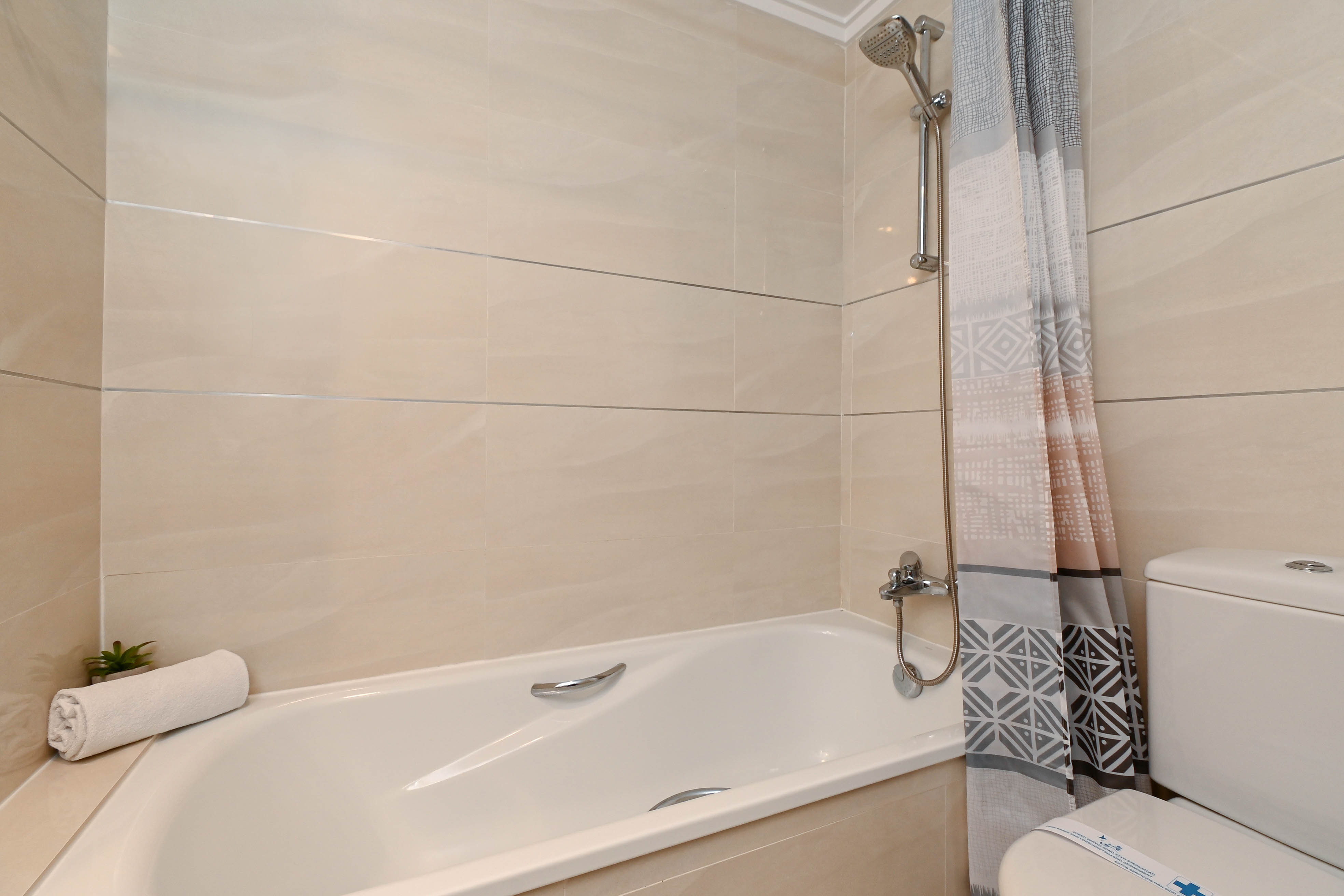 Functionele badkamer met ligbad, chromen kranen en beige wandtegels met horizontale accenten. Voorzien van een douchegordijn met geometrisch patroon.